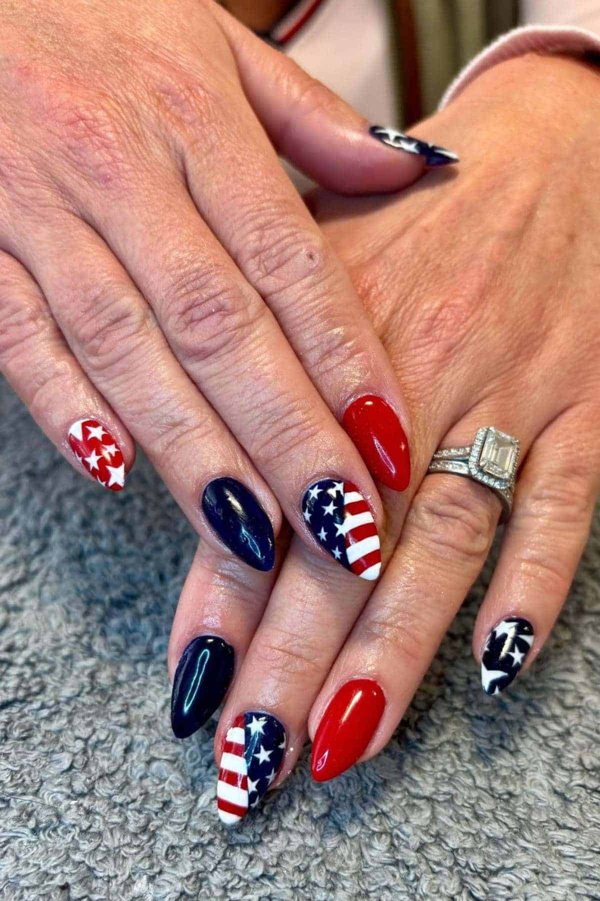 Close-up of hands with almond-shaped nails painted in red, white, and blue with American flag and star designs; person is wearing a diamond ring.