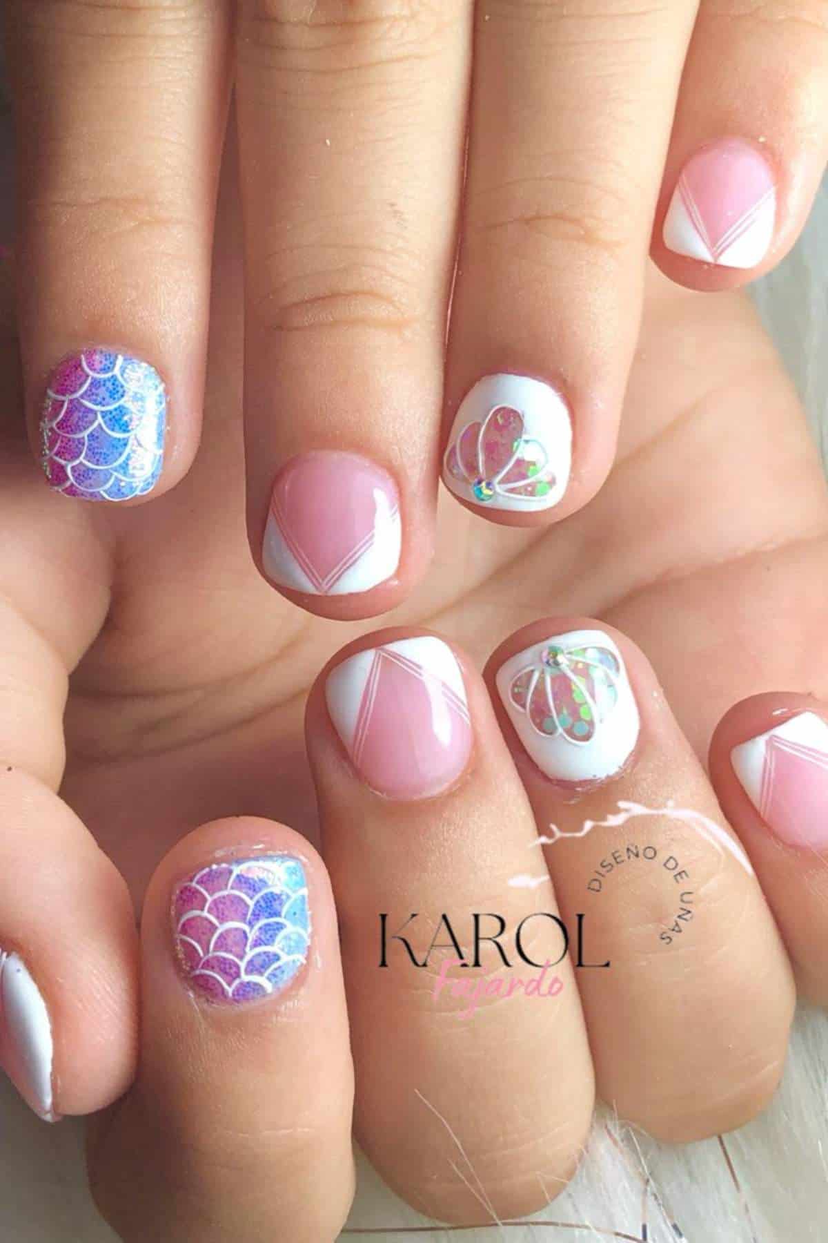 A hand with short nails featuring white geometric French tips and colorful fish scale and seashell designs on accent nails.