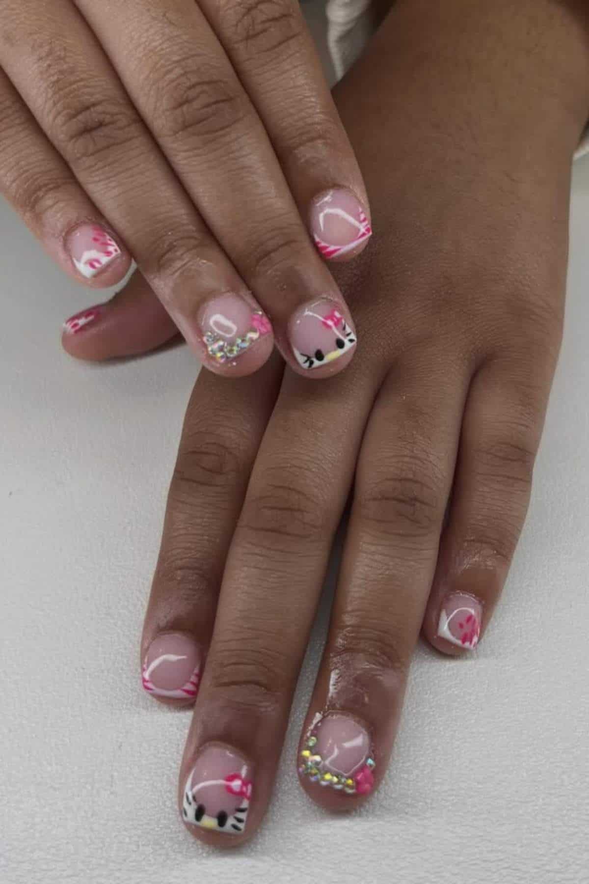 Hands with short, manicured nails featuring Hello Kitty designs, pink tips, and rhinestone accents on a white surface.