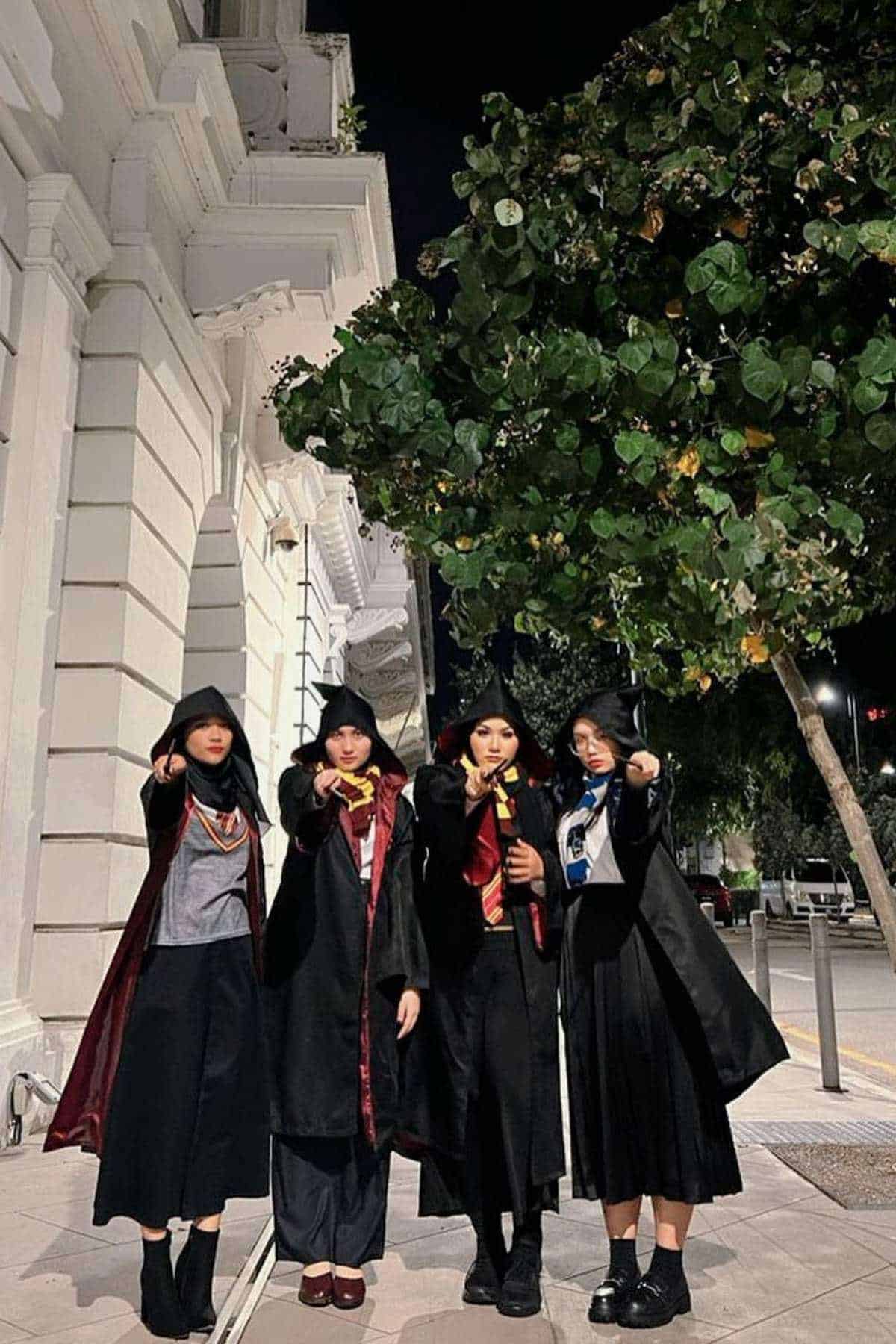 Four people stand on a city sidewalk at night, wearing robes and holding wands, resembling characters from the Harry Potter series.