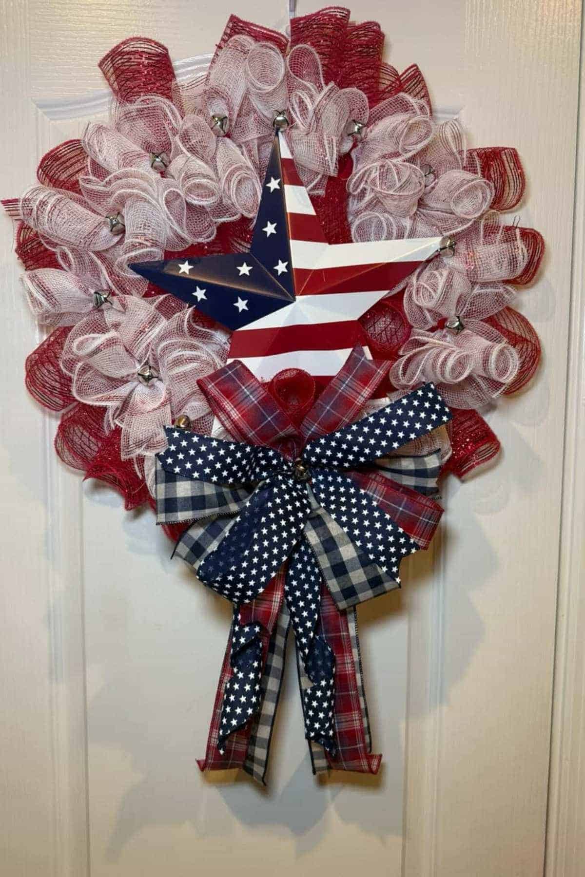 A patriotic wreath with red, white, and blue mesh, a star with stars and stripes, and a large bow featuring plaid and star patterns, hanging on a white door.