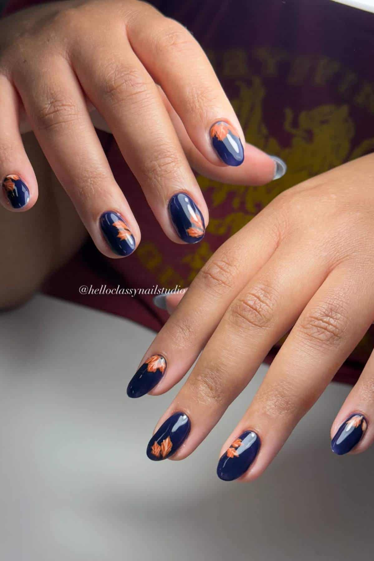 Hands with short, oval-shaped nails painted dark blue, each featuring a small orange leaf design near the cuticle.