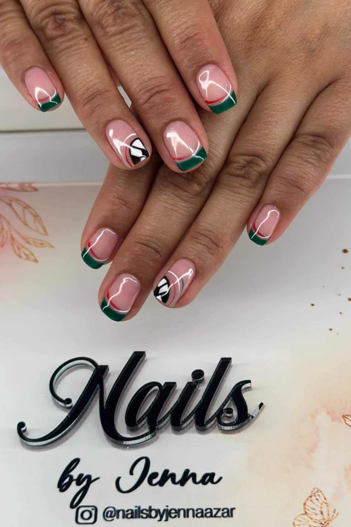Hands with manicured nails featuring green French tips and abstract white and black line art accents, displayed on a table with "Nails by Jenna" branding.
