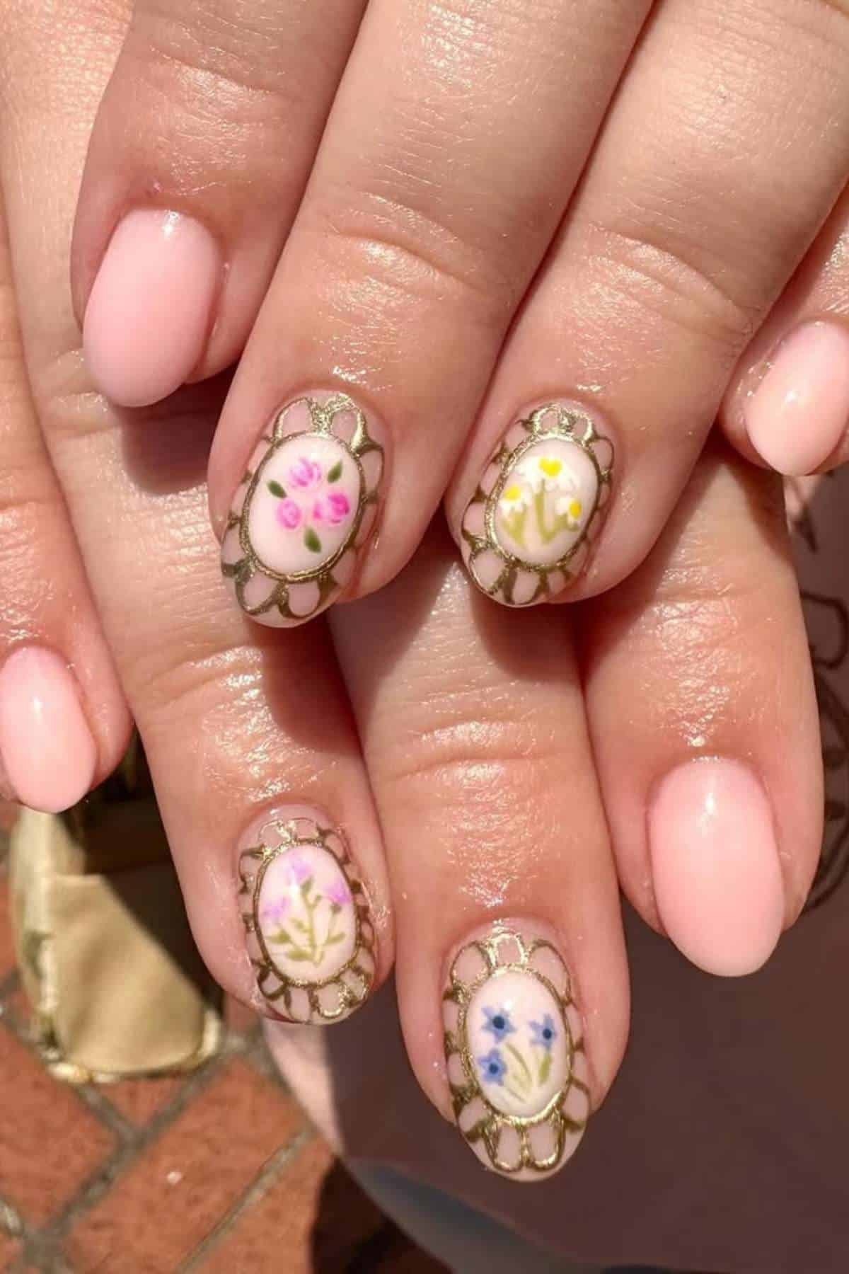 A close-up of hands with oval-shaped nails featuring pale pink polish and intricate floral designs framed by gold borders on four nails.