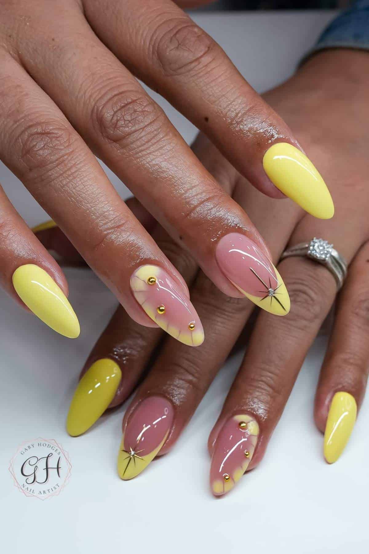 Hands with almond-shaped nails featuring yellow tips, gold bead accents, and starburst designs on a few nude-pink base nails. One hand has a ring with clear stones.