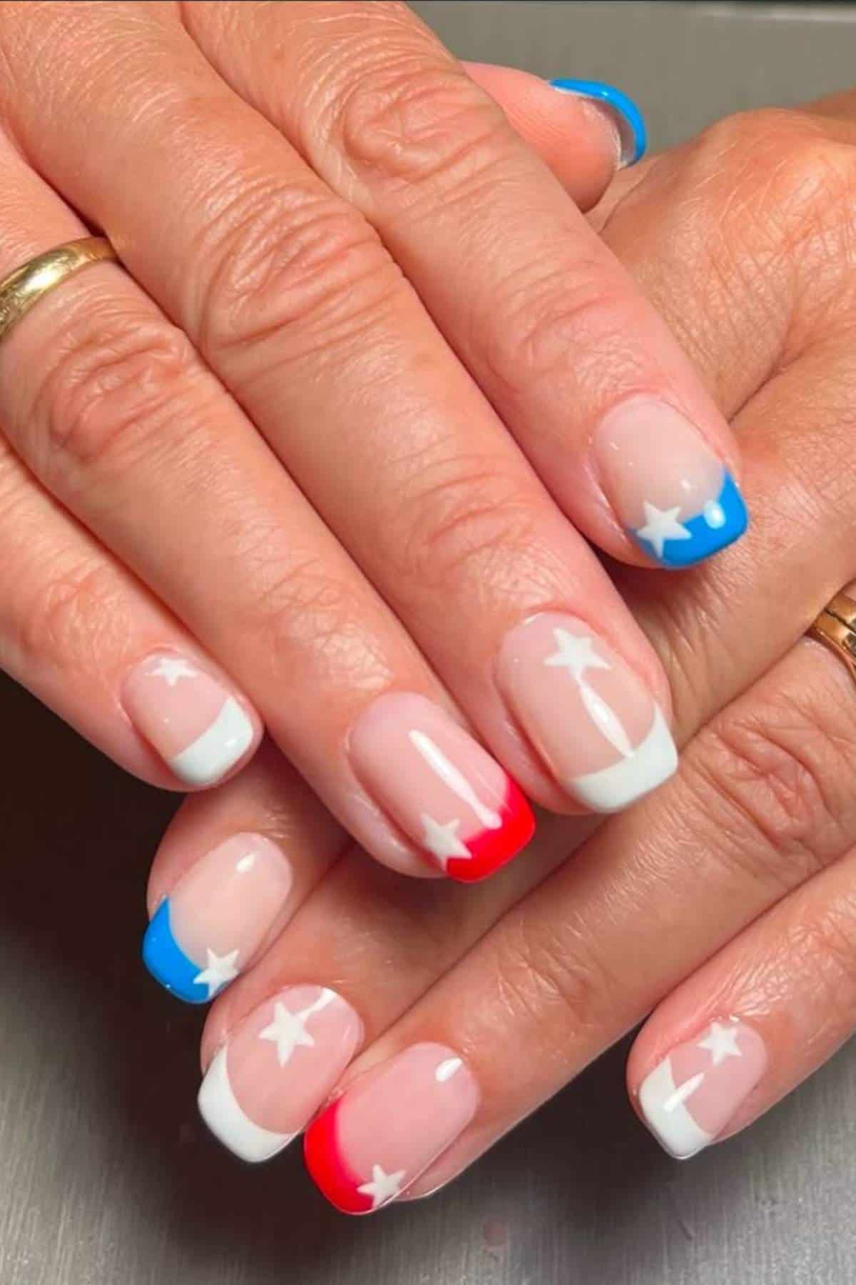 Close-up of hands with manicured nails featuring blue, red, and white French tips and white star designs, inspired by the colors of the Texas flag.