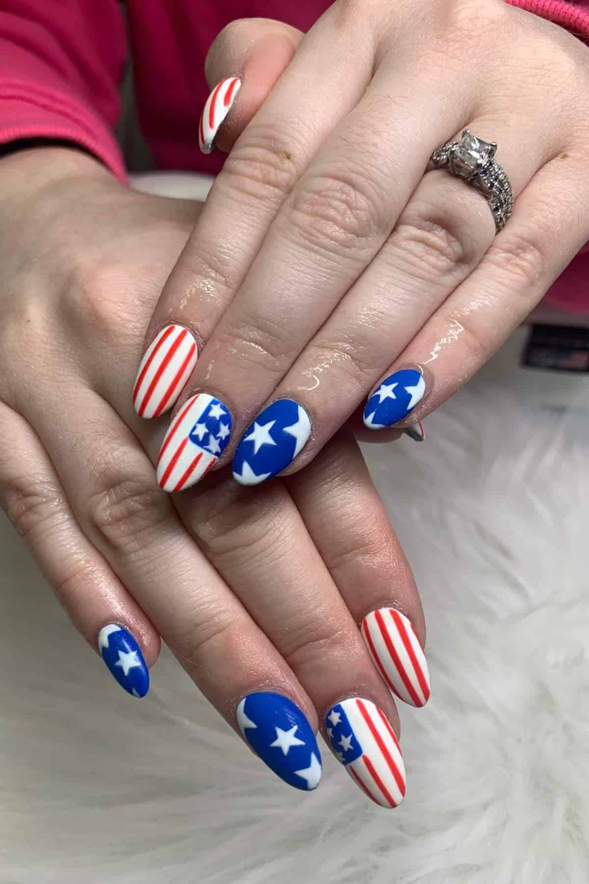 Hands with long, almond-shaped nails painted with red and white stripes and blue backgrounds with white stars, resembling the American flag.