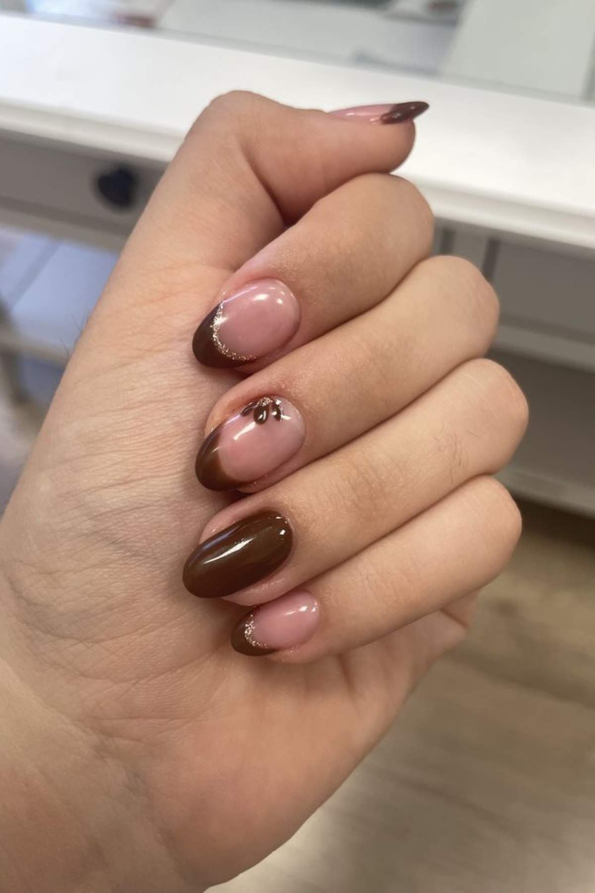 A hand with almond-shaped nails featuring a brown French tip design, nude bases, gold glitter accents, and a small gold embellishment on the ring finger.