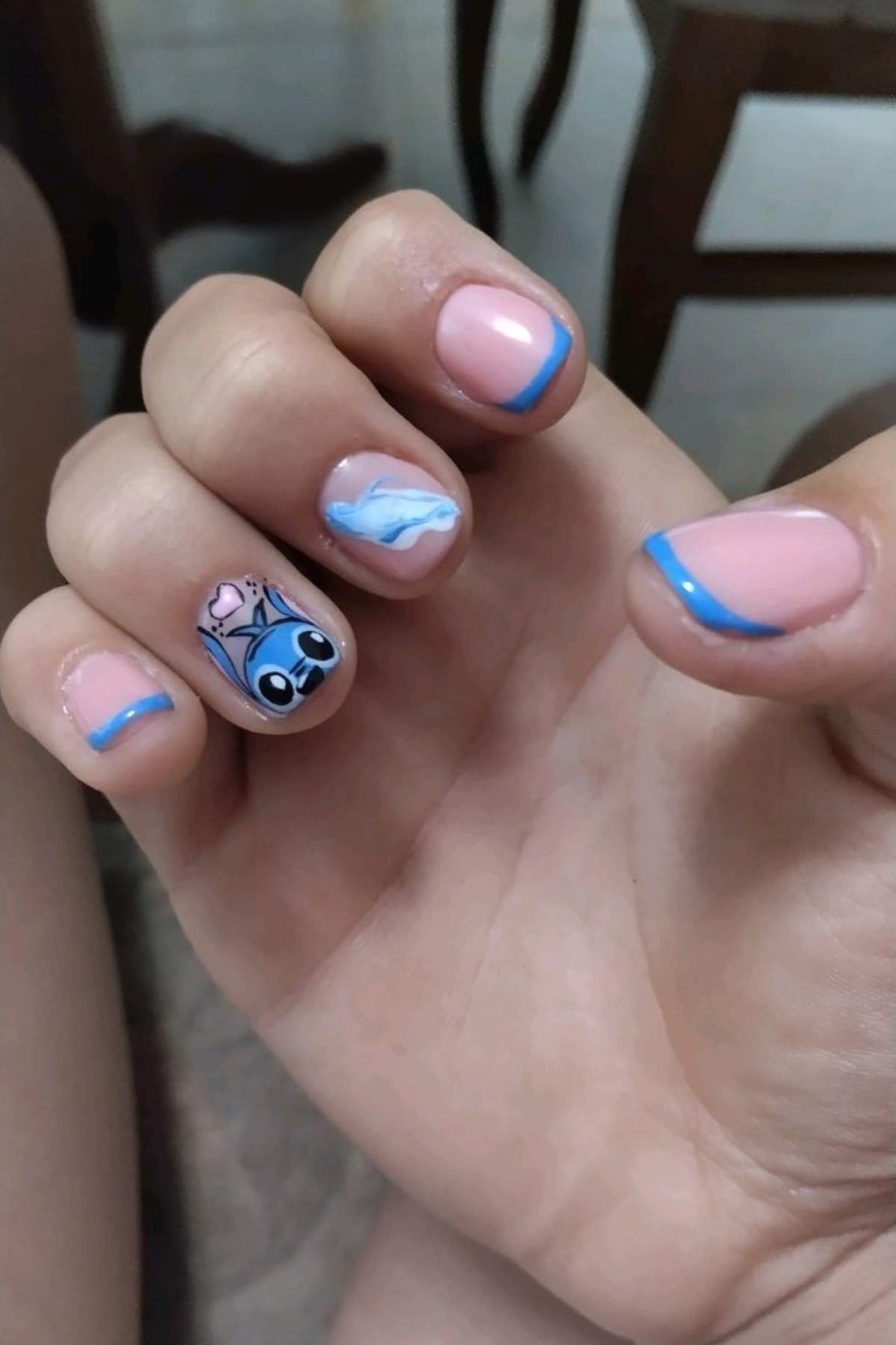 A hand with pink nails, each featuring a thin blue tip, and one nail decorated with a cartoon character and wave design.