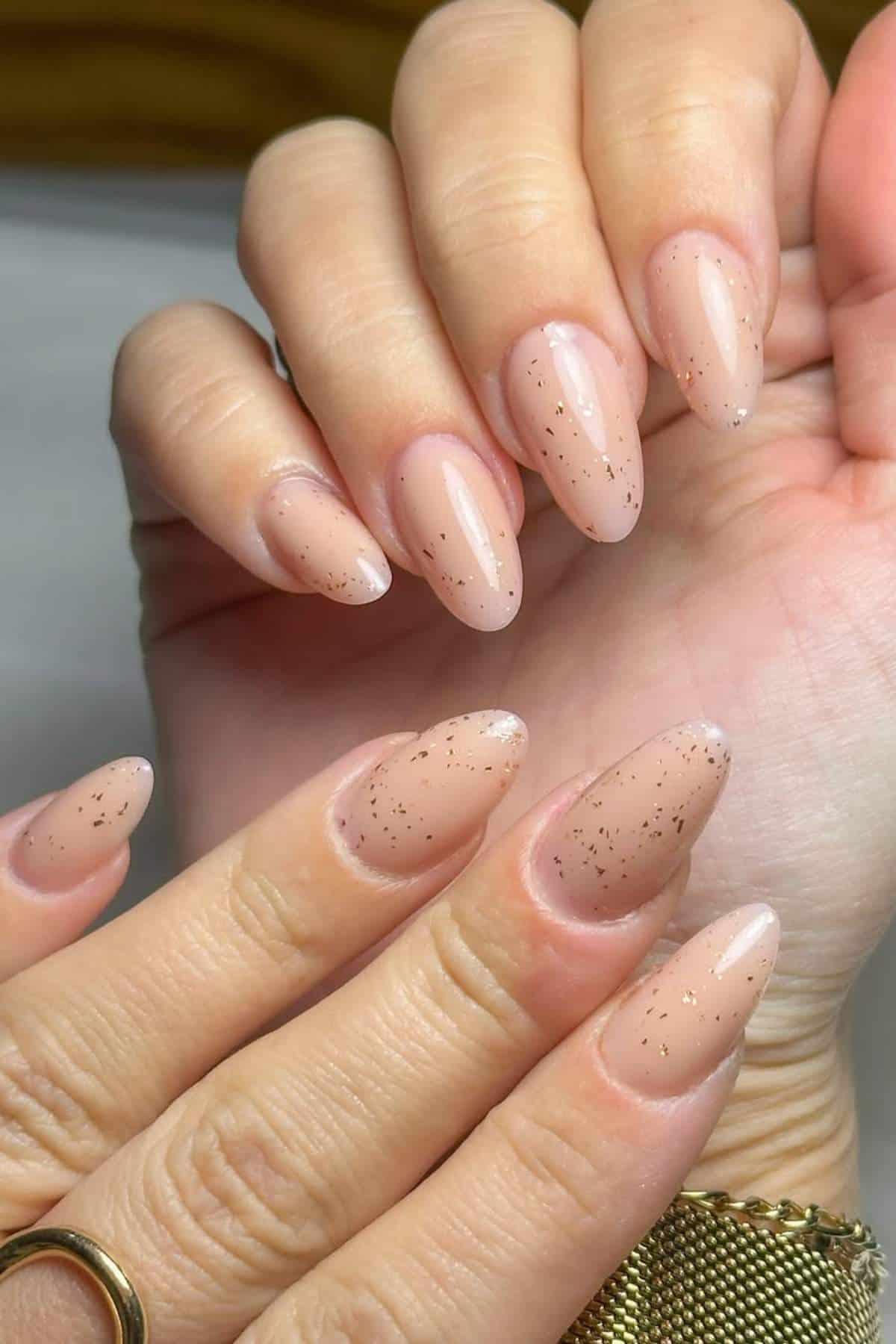 Close-up of hands with almond-shaped nails painted in a nude color with subtle gold glitter accents.