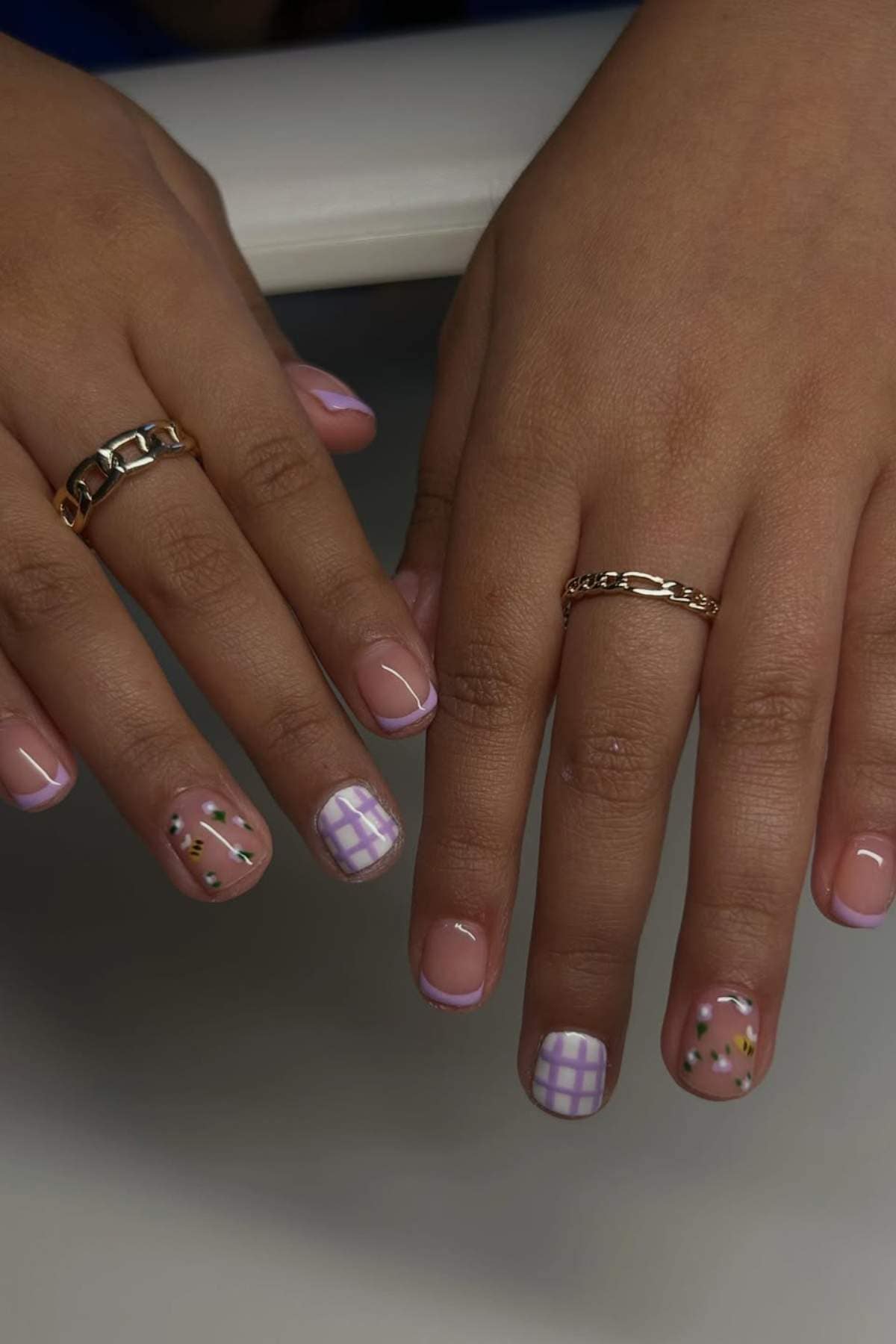 Two hands with gold rings display manicured nails featuring pink French tips, purple grid patterns, and floral designs with gold accents.