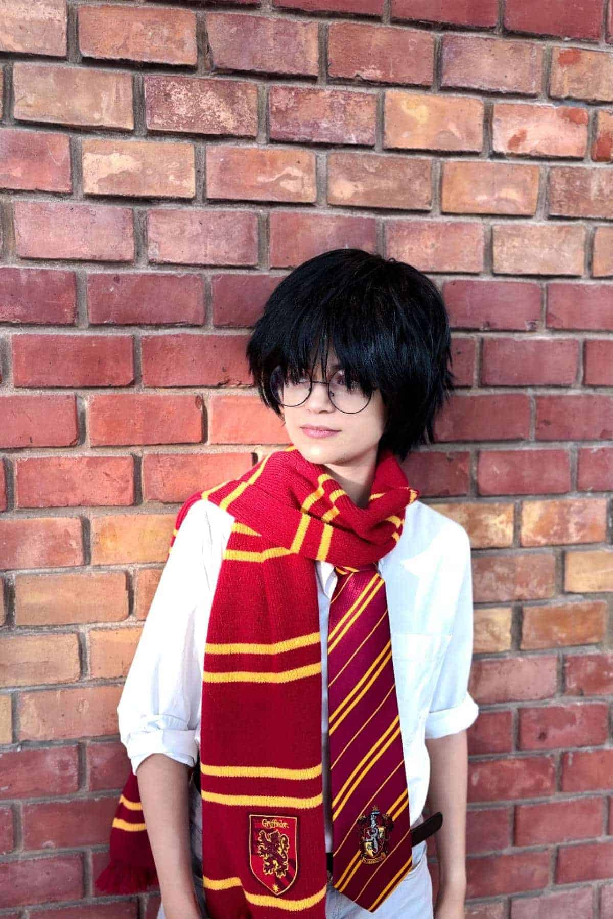 Person wearing round glasses, a white shirt, and a red-and-gold striped scarf and tie, standing against a brick wall.
