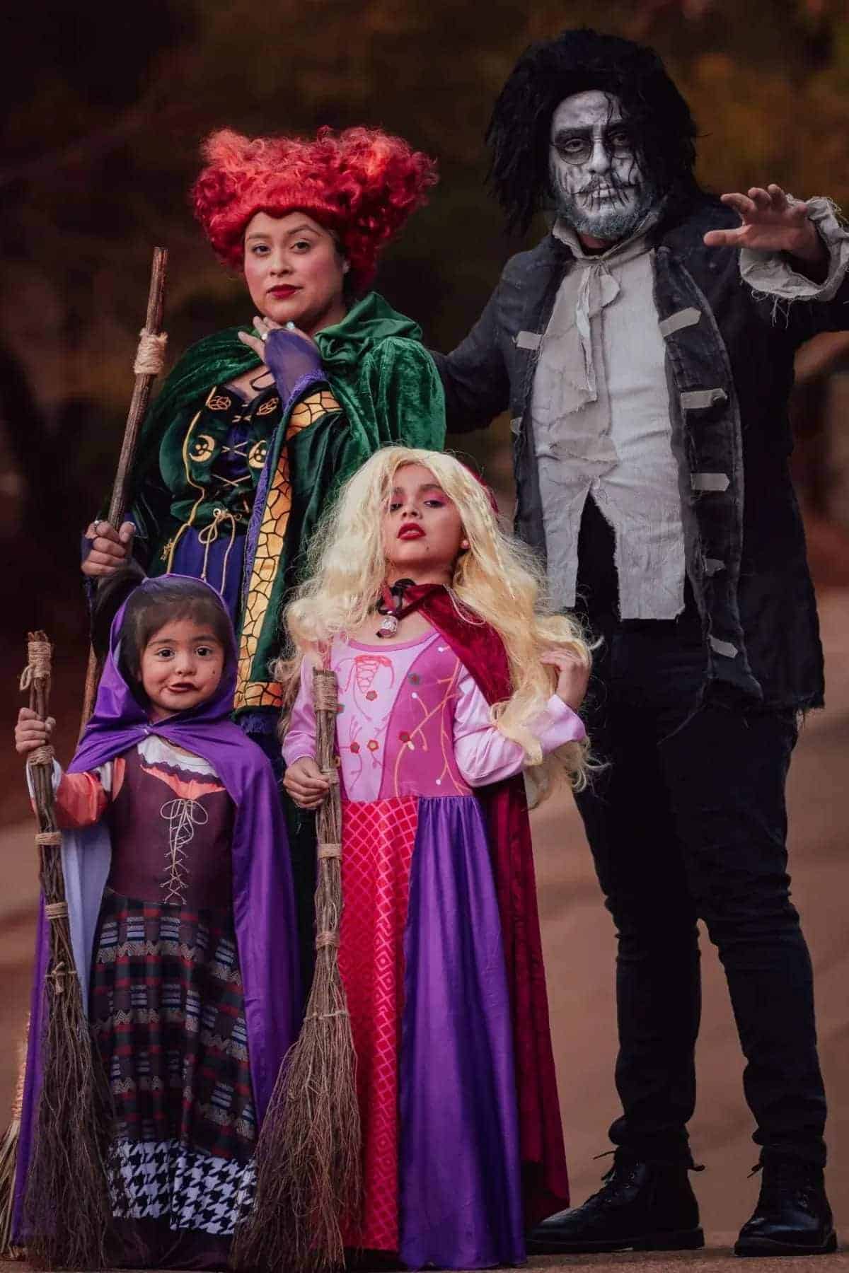 Four people dressed in elaborate Halloween costumes pose together, with two adults and two children holding brooms and wearing colorful wigs and robes.
