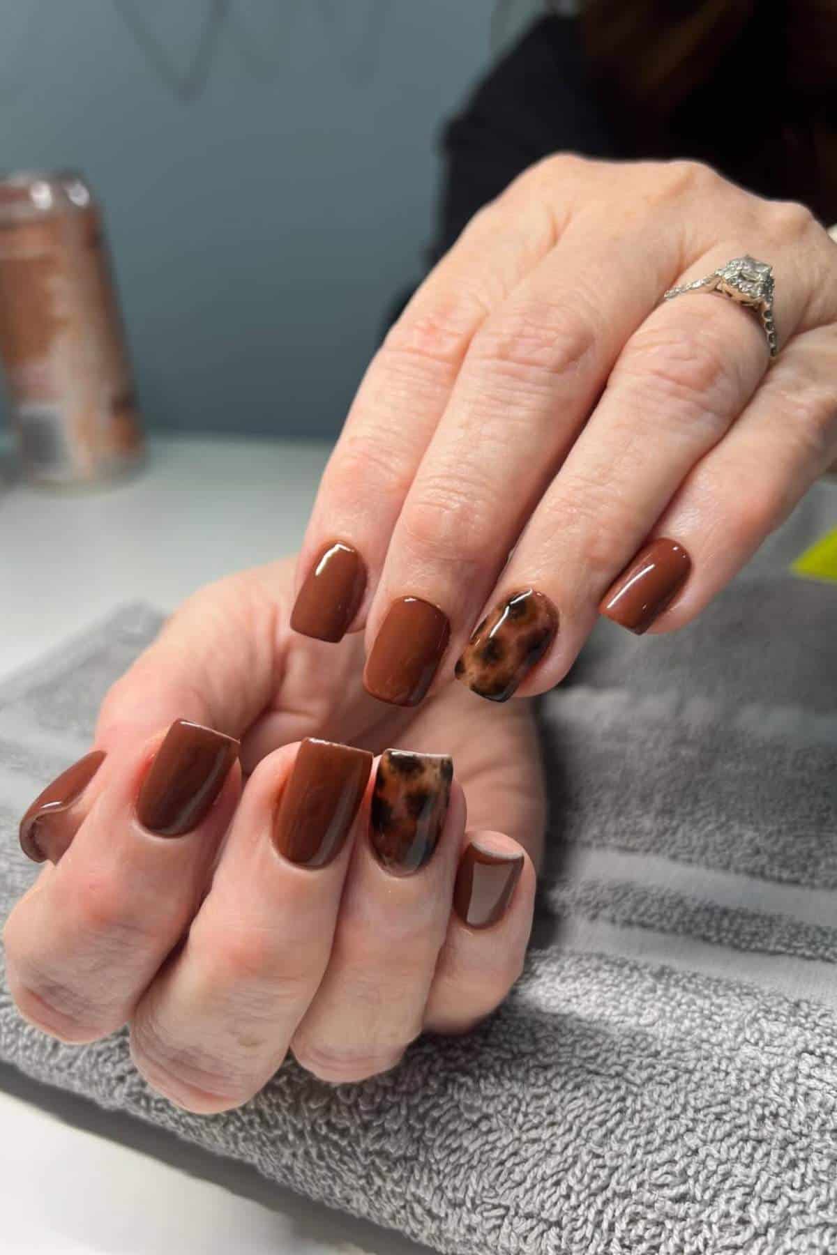 A person displays freshly manicured nails painted brown, with one accent nail featuring a tortoiseshell pattern on each hand.