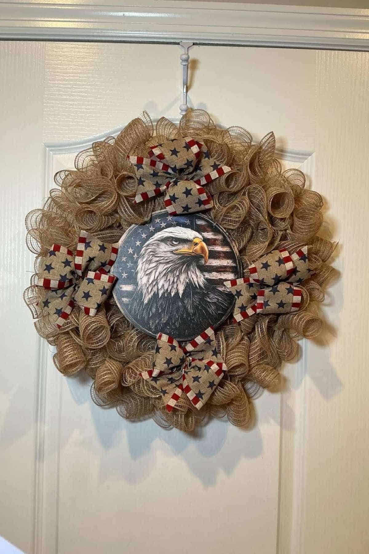 A decorative wreath with burlap loops, star-patterned bows, and a centerpiece featuring a bald eagle with an American flag background, hanging on a white door.