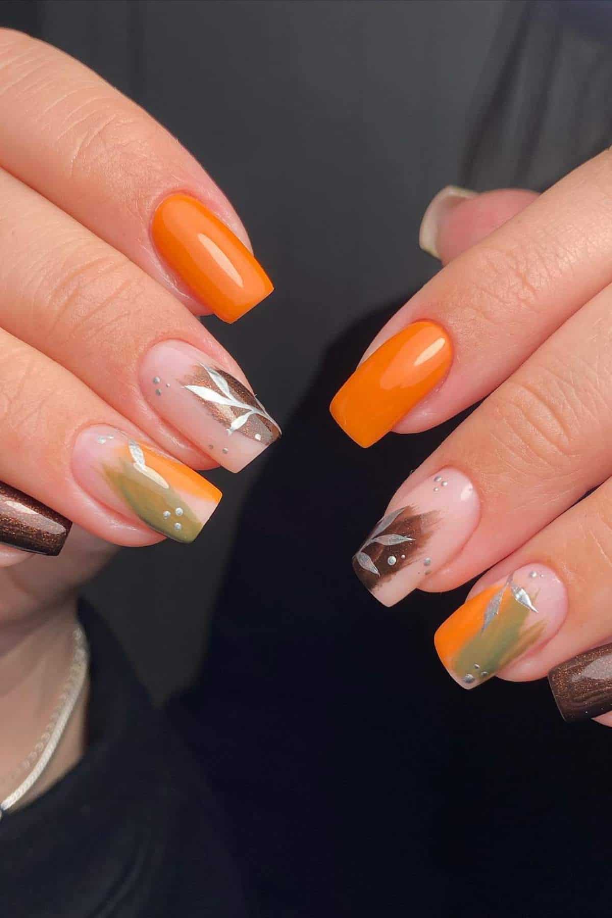 Hands with square-shaped nails featuring orange, brown, and olive green nail art, including solid colors, abstract designs, and silver dot accents.