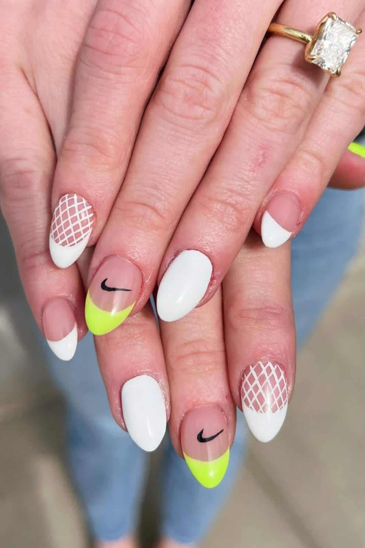 Hands with almond-shaped nails featuring white polish, neon yellow tips, black Nike swoosh logos, and white net designs on some nails. A gold ring with a large rectangular stone is worn on one finger.