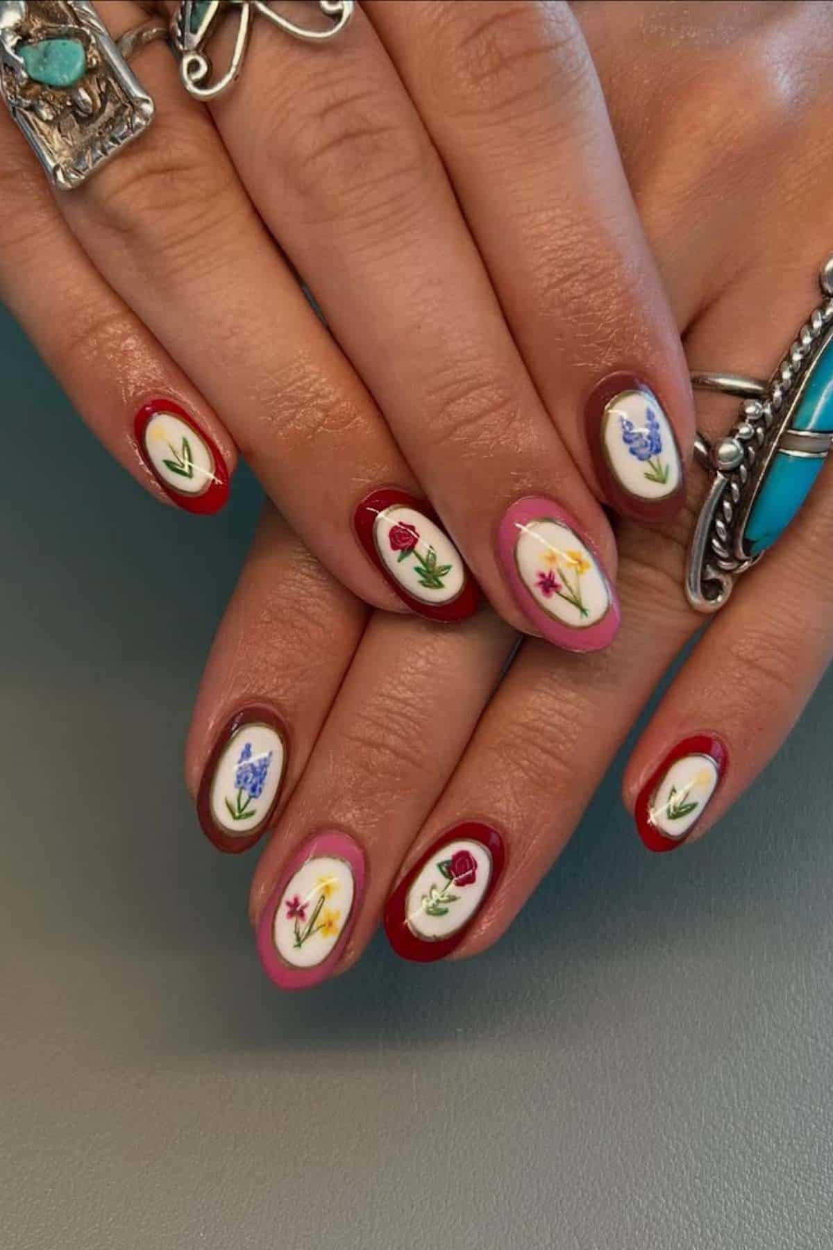 A hand with oval-shaped nails painted with floral designs and red, pink, or brown borders; person is wearing silver rings with turquoise stones.