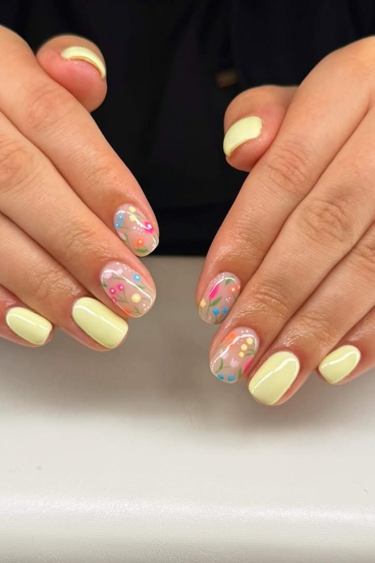 Hands with light yellow nail polish; some nails feature clear polish with colorful floral designs.