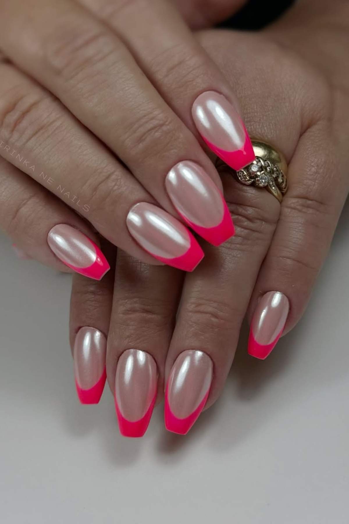 Hands with coffin-shaped nails featuring a pearly base color and bright pink French tips. The person is wearing gold rings on their fingers.