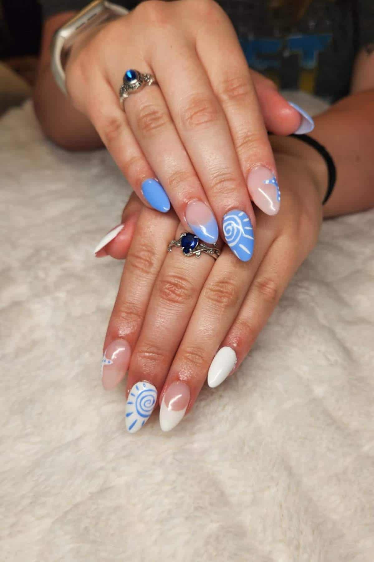 Hands with long, almond-shaped nails painted in blue, white, and nude, featuring swirl and shell designs, resting on a white textured surface.