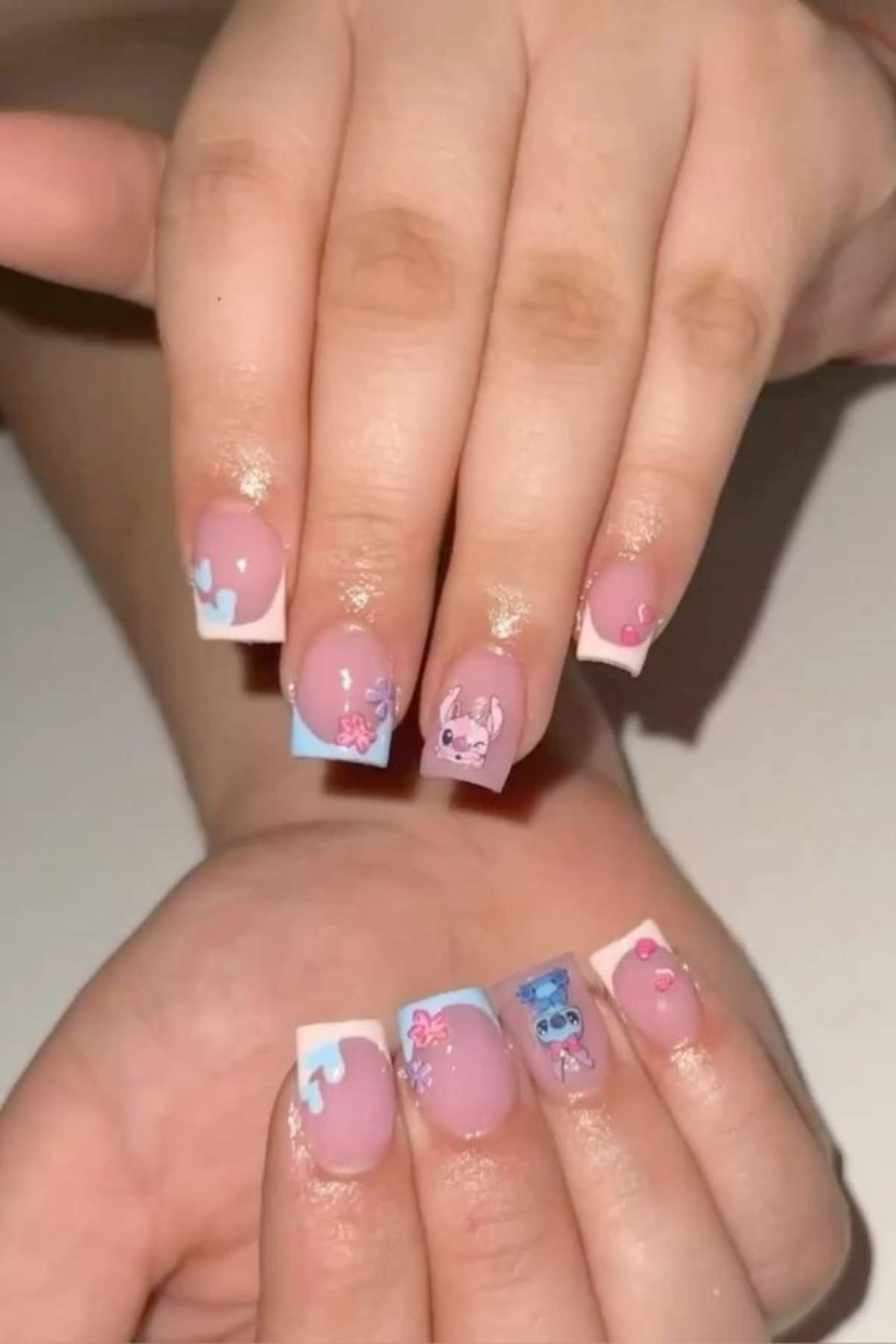 Hands with long, square-shaped nails featuring pastel pink and blue French tips, flower designs, and cartoon character nail art.