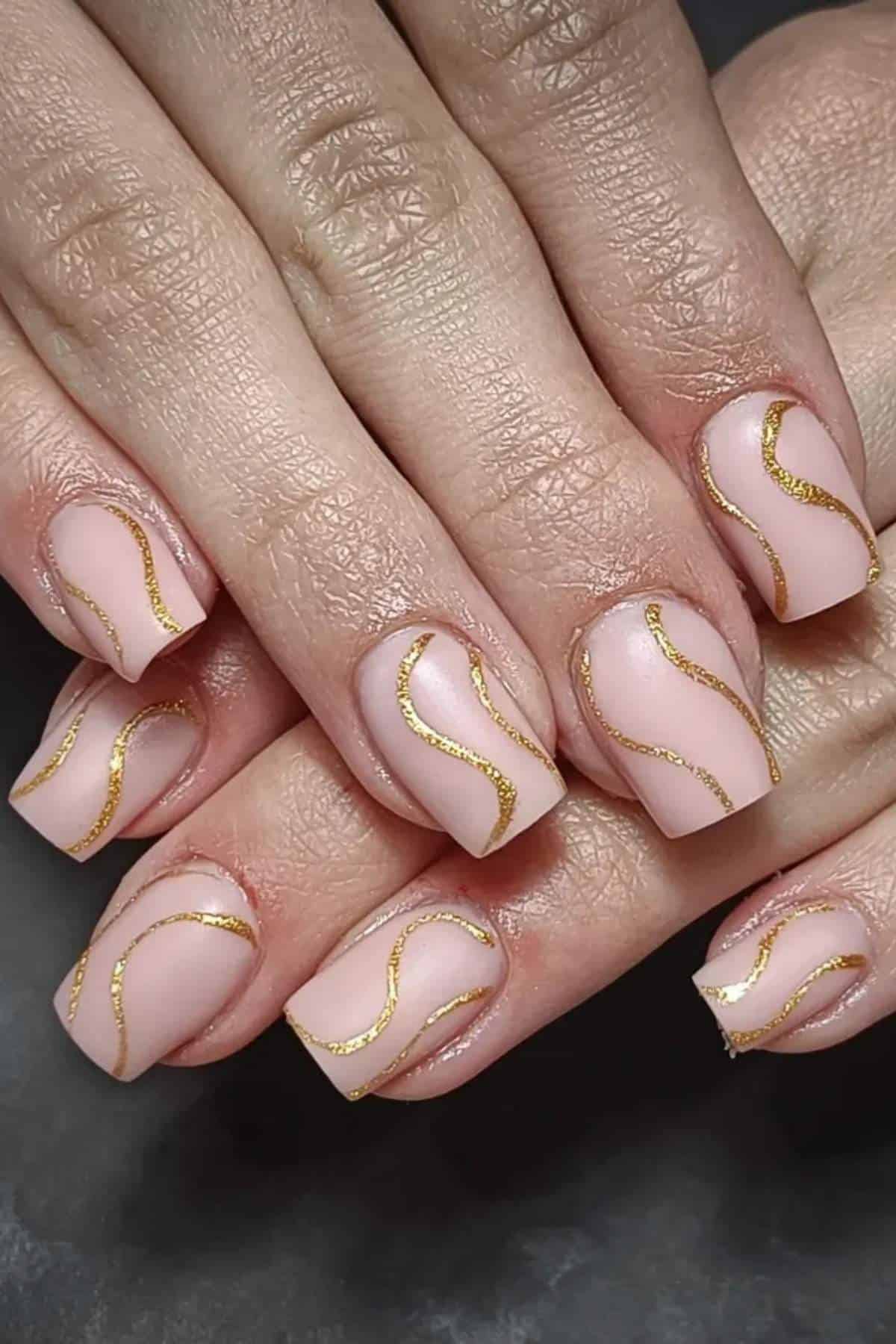 Close-up of hands with short square nude nails featuring gold wavy line designs on each nail.