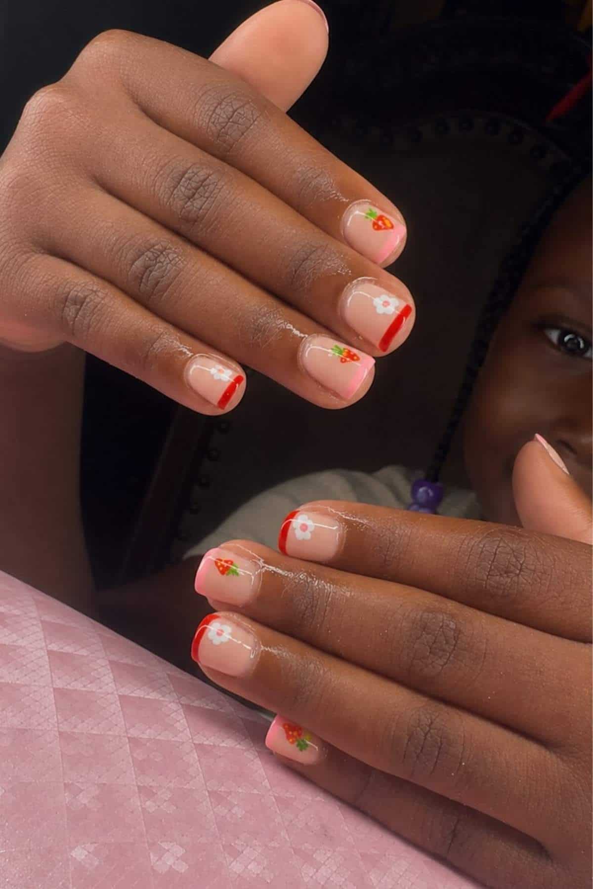 Close-up of hands with natural nails featuring red tips and small floral designs, with a child smiling in the background.