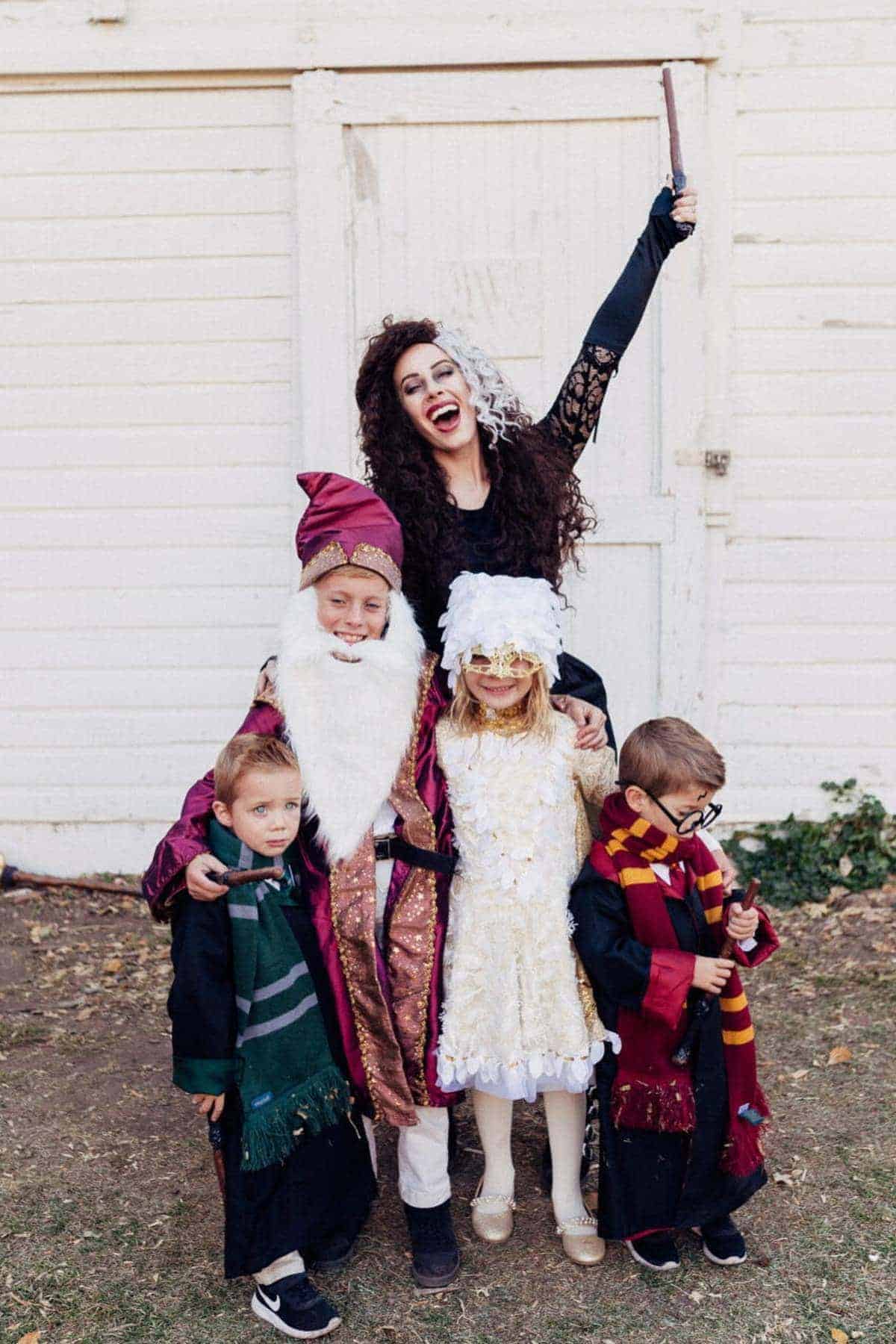 A woman dressed as Bellatrix Lestrange poses with four children in Harry Potter costumes, standing outside in front of a white wooden building.