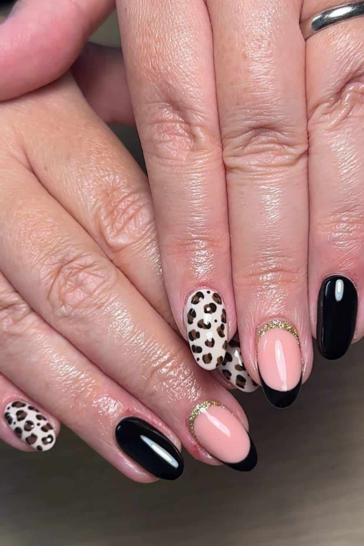 Hands with medium-length almond-shaped nails featuring black polish, nude polish with gold accents, and an animal print design on two nails.