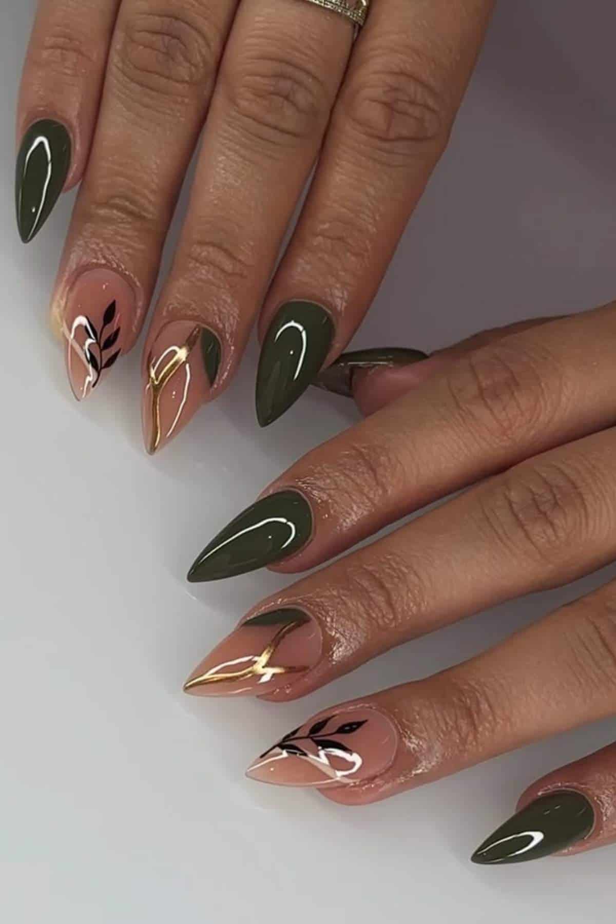 Hands with long, almond-shaped nails featuring olive green, nude, and gold colors, with black leaf designs and glossy finish.