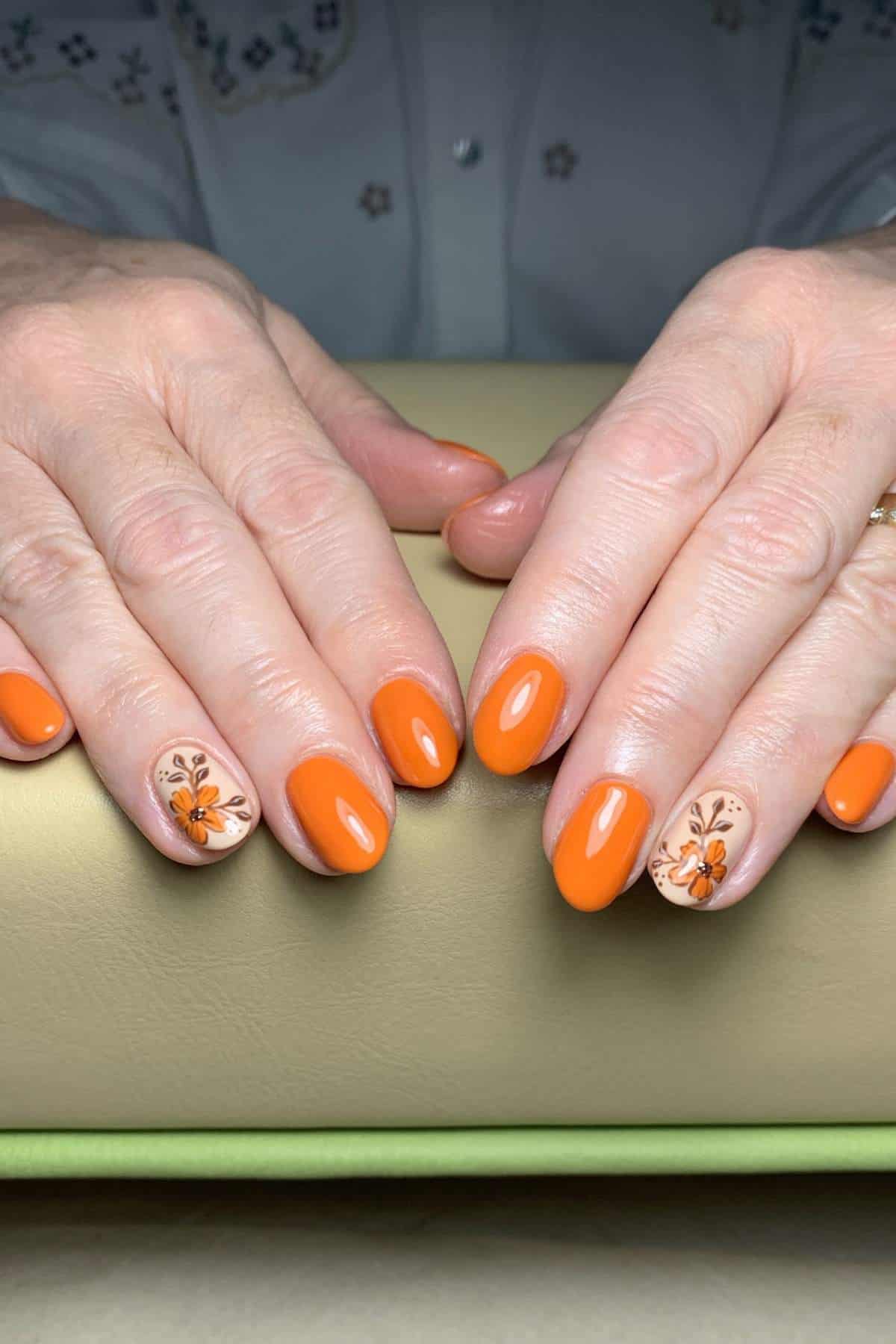 Hands with orange manicured nails; ring fingers feature a floral and butterfly design on a neutral base.