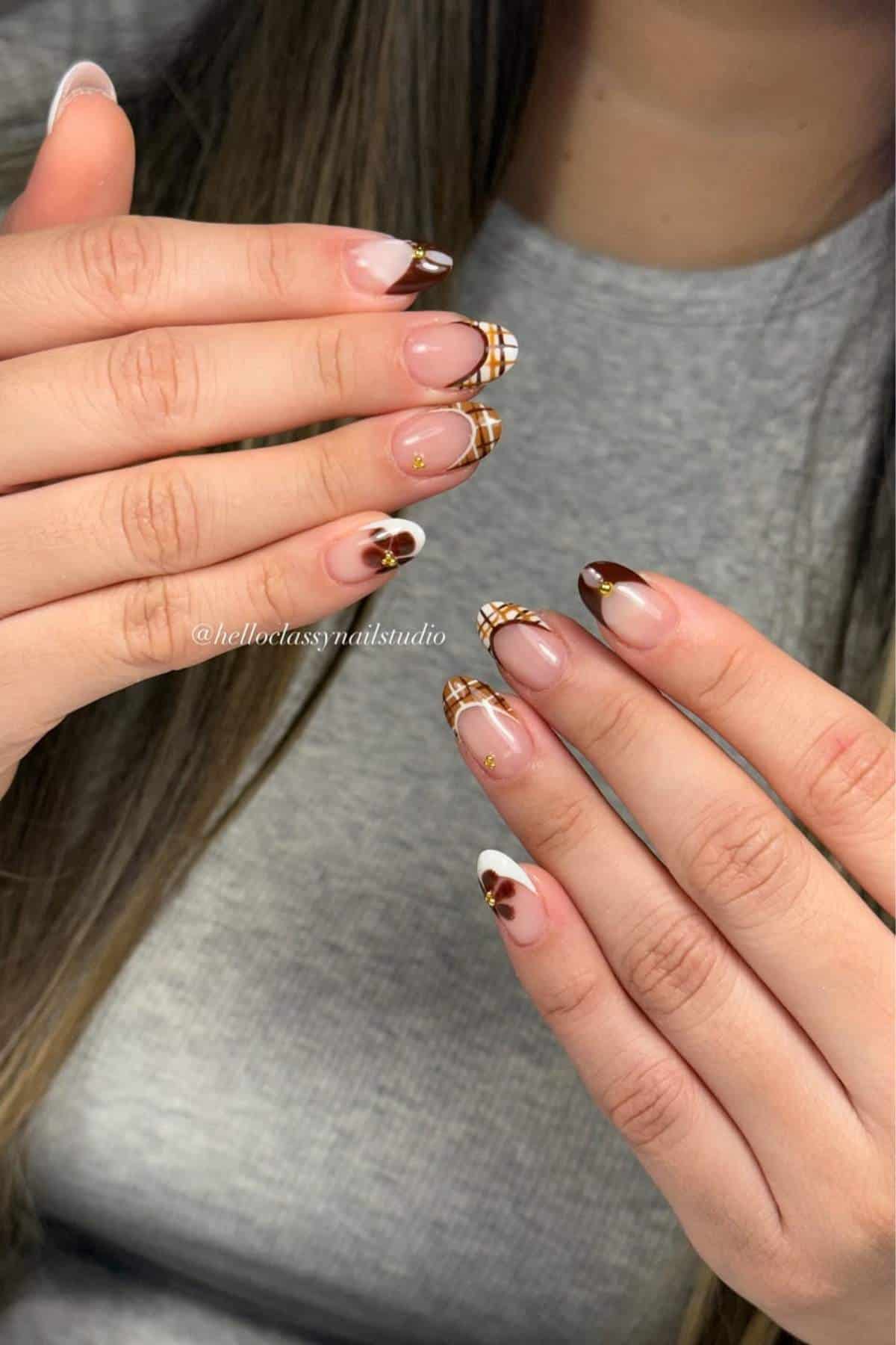 Close-up of hands with long almond-shaped nails featuring brown, white, and gold plaid and heart nail art designs. Person is wearing a grey top.