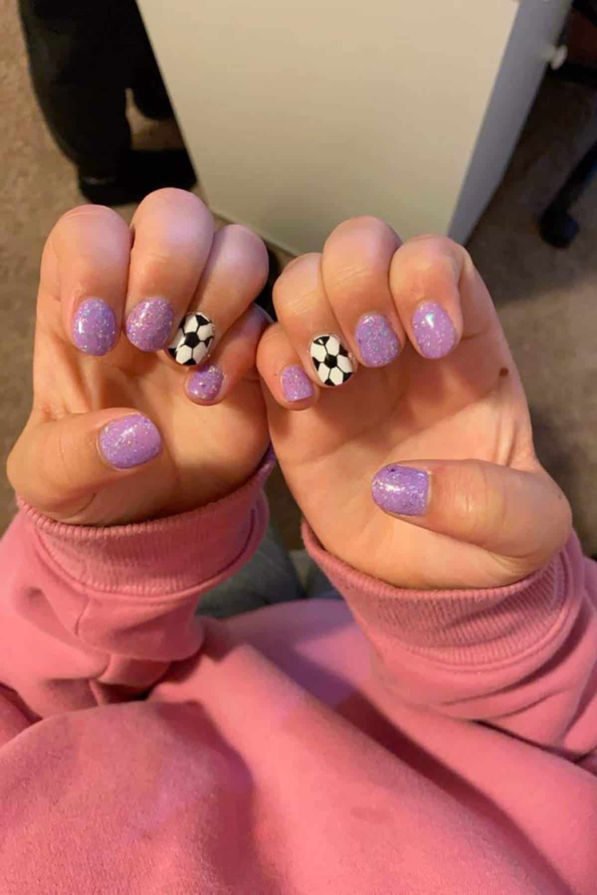 Two hands showing sparkly purple nail polish with an accent nail on each hand featuring a black and white soccer ball design.