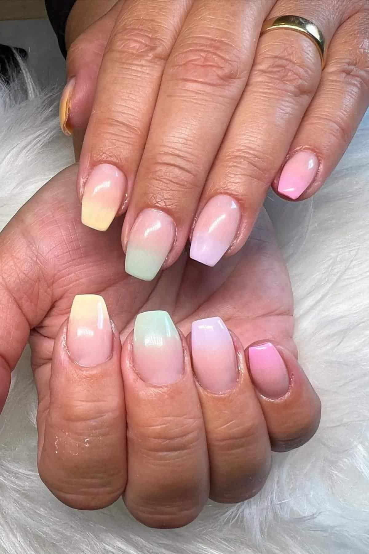 Close-up of hands with short, ballerina-shaped nails painted in pastel gradient colors including yellow, green, purple, and pink.