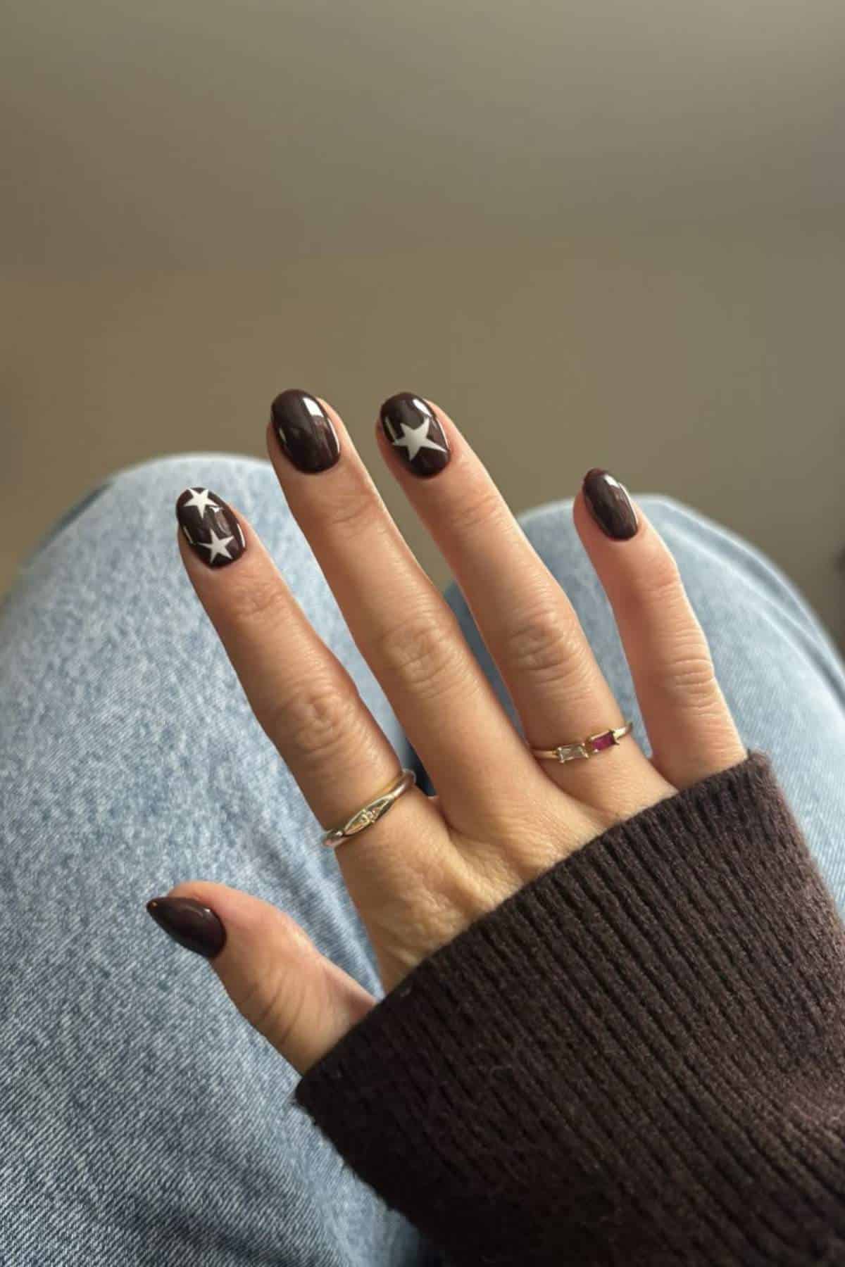 A hand with dark brown manicure featuring white star designs on two nails, wearing two rings, rests on denim-clad legs and a dark sweater sleeve.