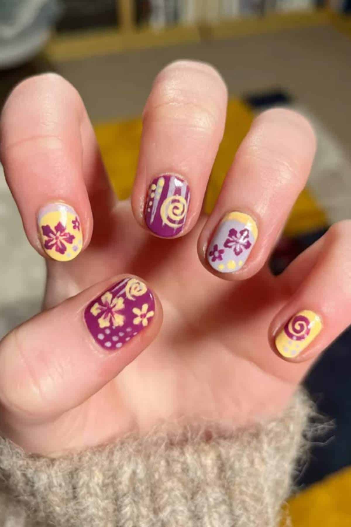 A hand with short nails painted in purple and yellow, featuring floral and swirl designs on each nail.