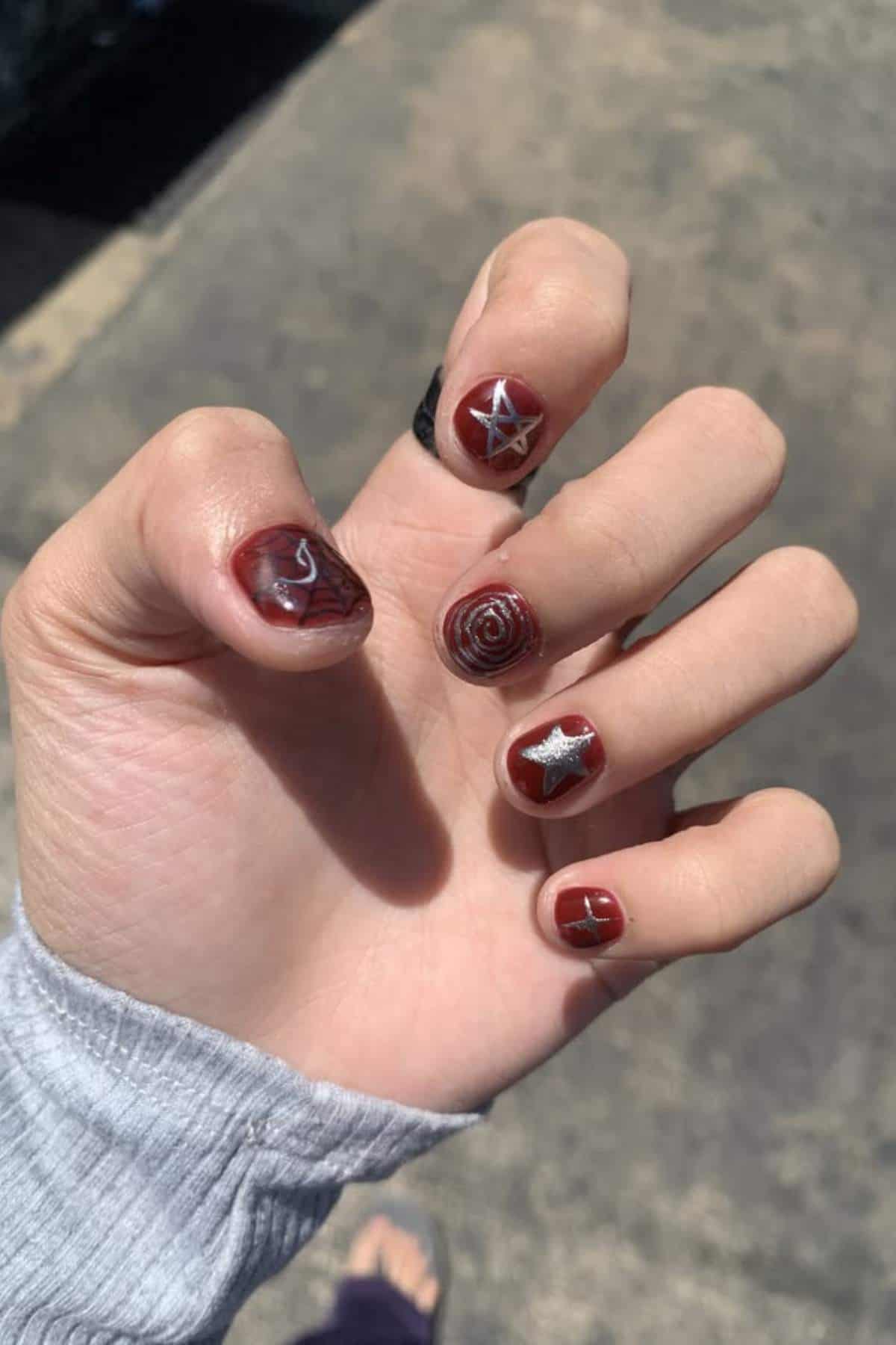 A hand with short nails painted dark red featuring abstract white and silver line designs, photographed outdoors in natural light.