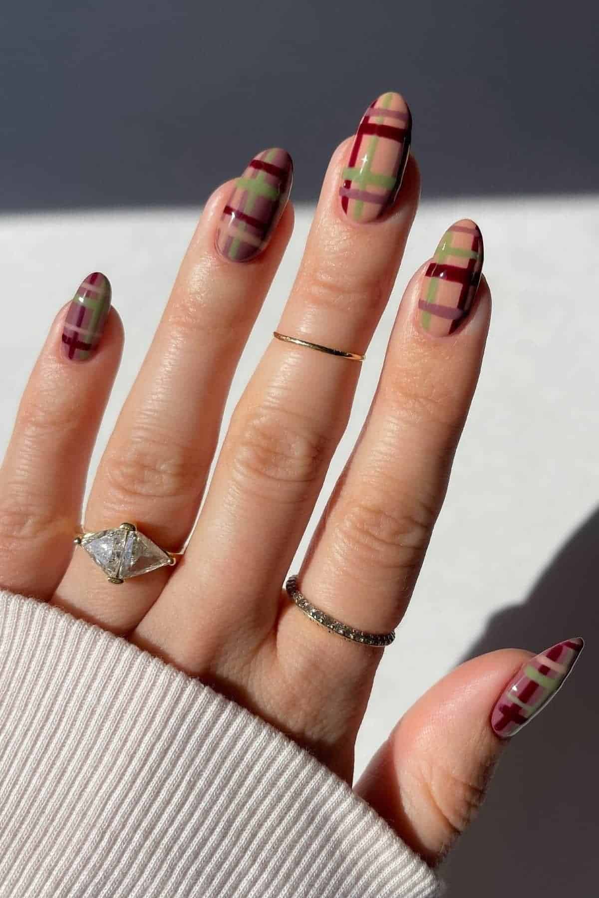 A hand with almond-shaped nails featuring a plaid pattern in burgundy, green, and beige. The person is wearing three rings and a cream-colored sleeve.