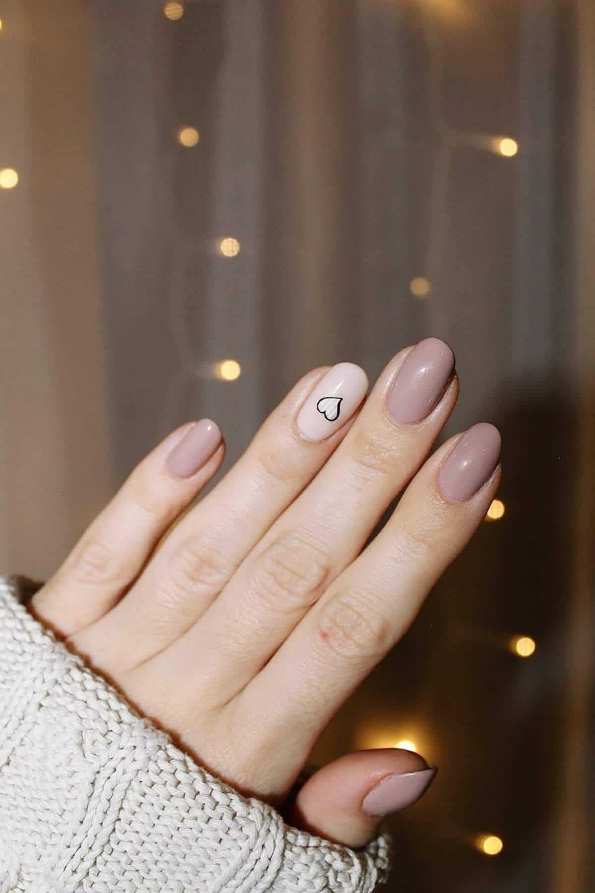 A hand with short, almond-shaped nails painted in nude polish, with the ring finger featuring a small black heart outline on a light accent nail.