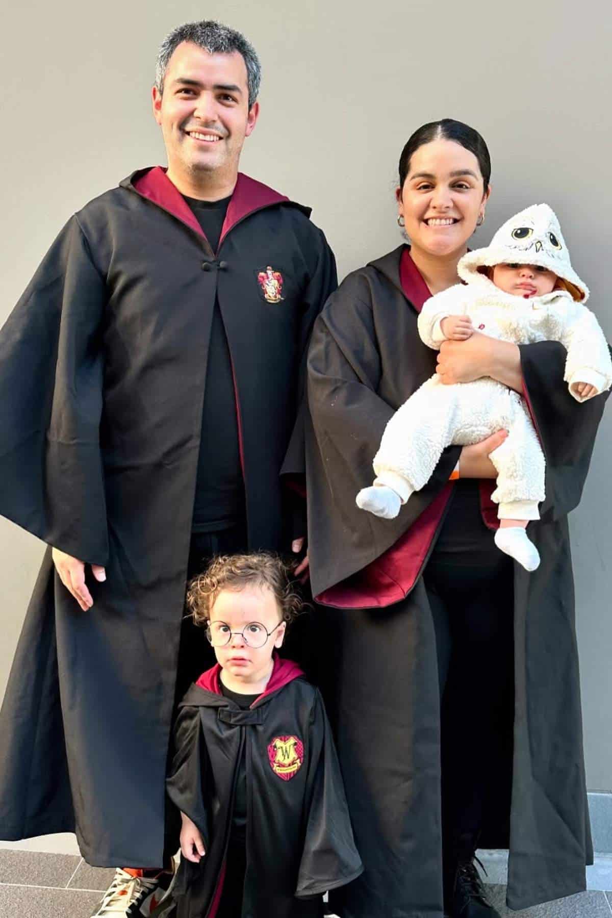 A family of four poses in Harry Potter-themed costumes; two adults and a child wear Hogwarts robes, while a baby is dressed in a white animal onesie.