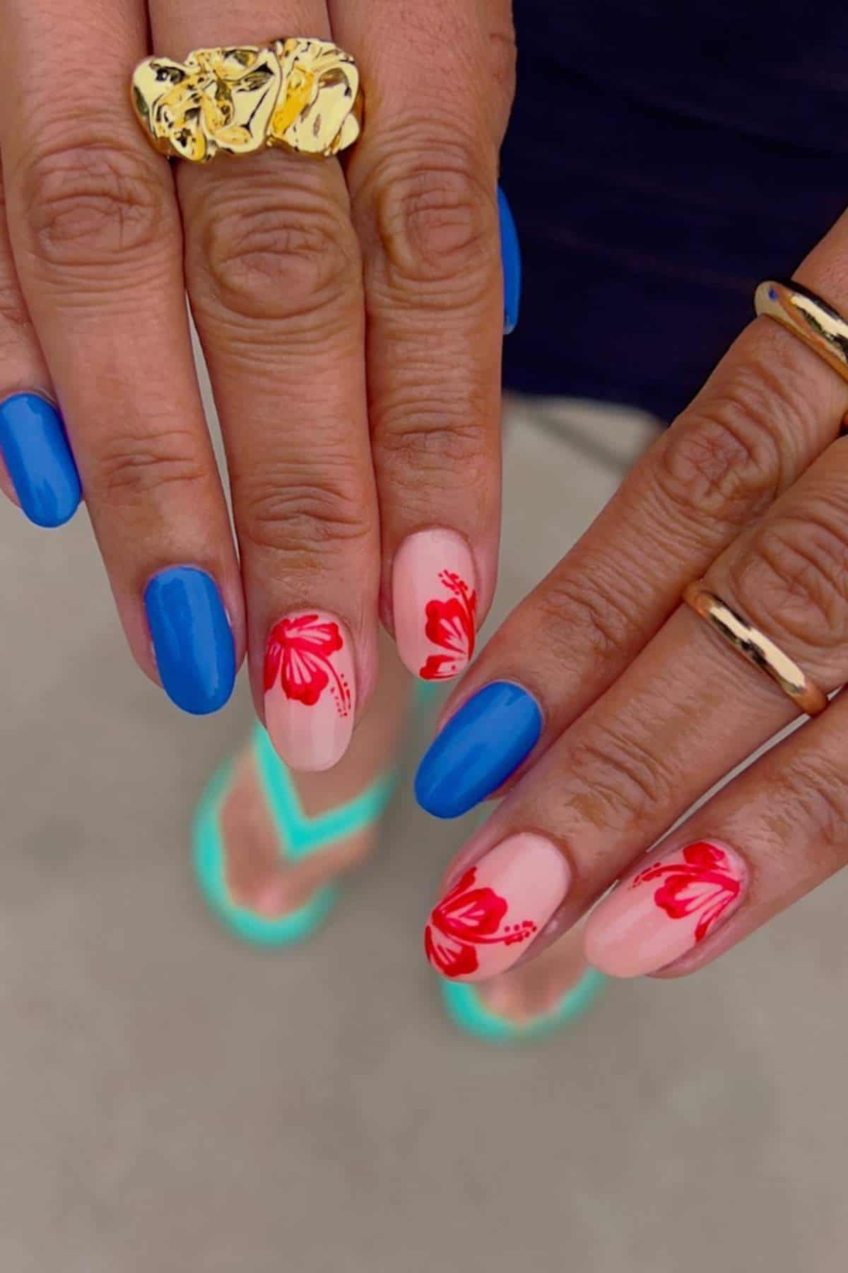 Close-up of hands with blue nail polish and light pink nails featuring red floral designs, accessorized with gold rings; person is wearing teal sandals.