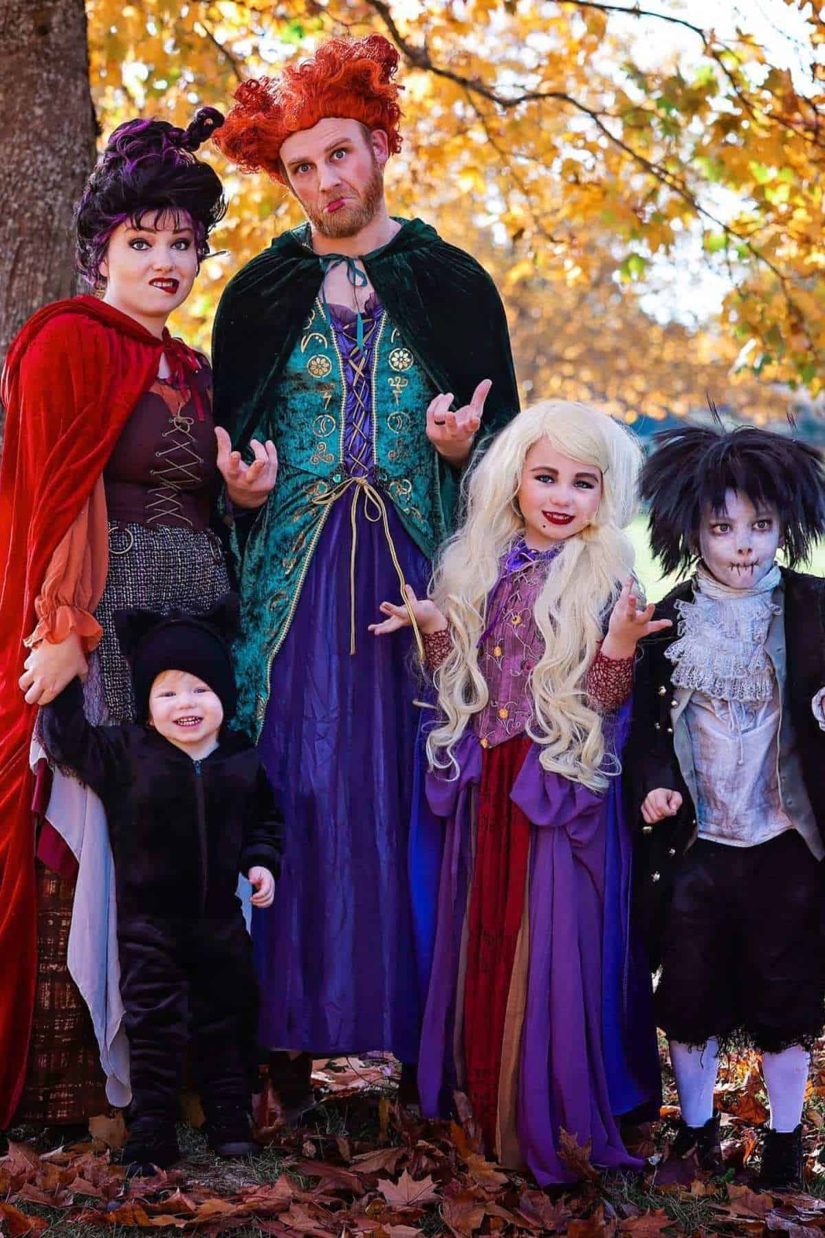 A group of five people, including children, are dressed in colorful Halloween costumes resembling witches and a vampire, standing outdoors with autumn leaves in the background.