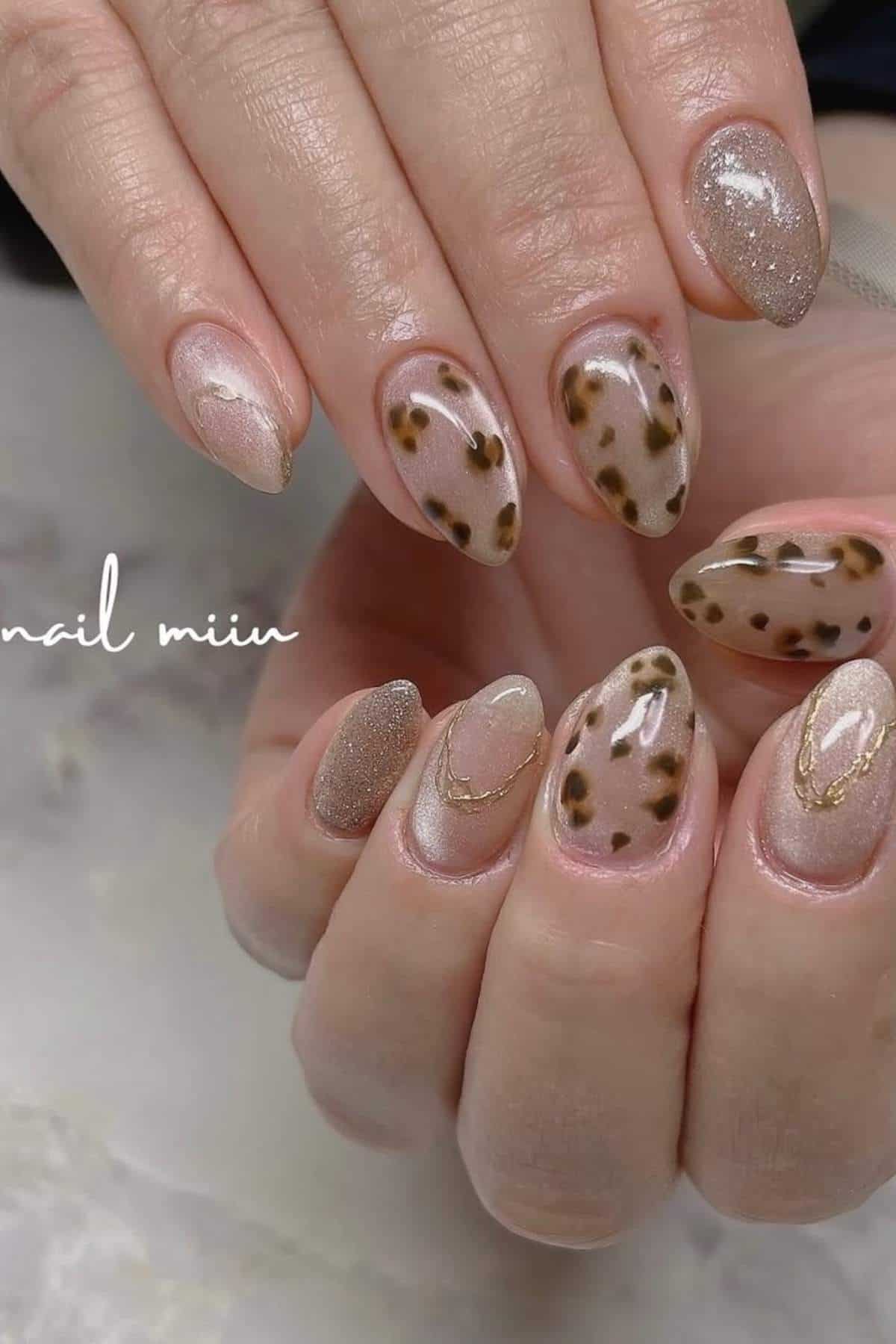 Hands with almond-shaped nails featuring a mix of leopard print, nude, and glittery beige designs with subtle gold accents.