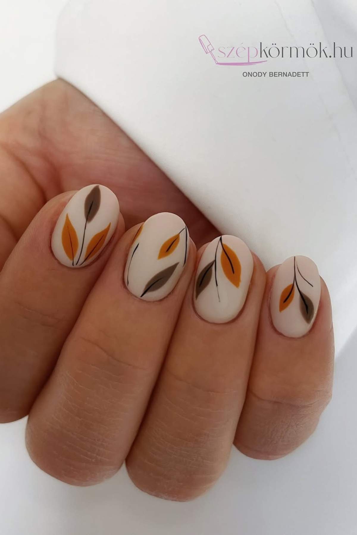 Close-up of a hand with short, almond-shaped nails featuring a matte nude base and minimalist leaf designs in brown, orange, and dark gray.