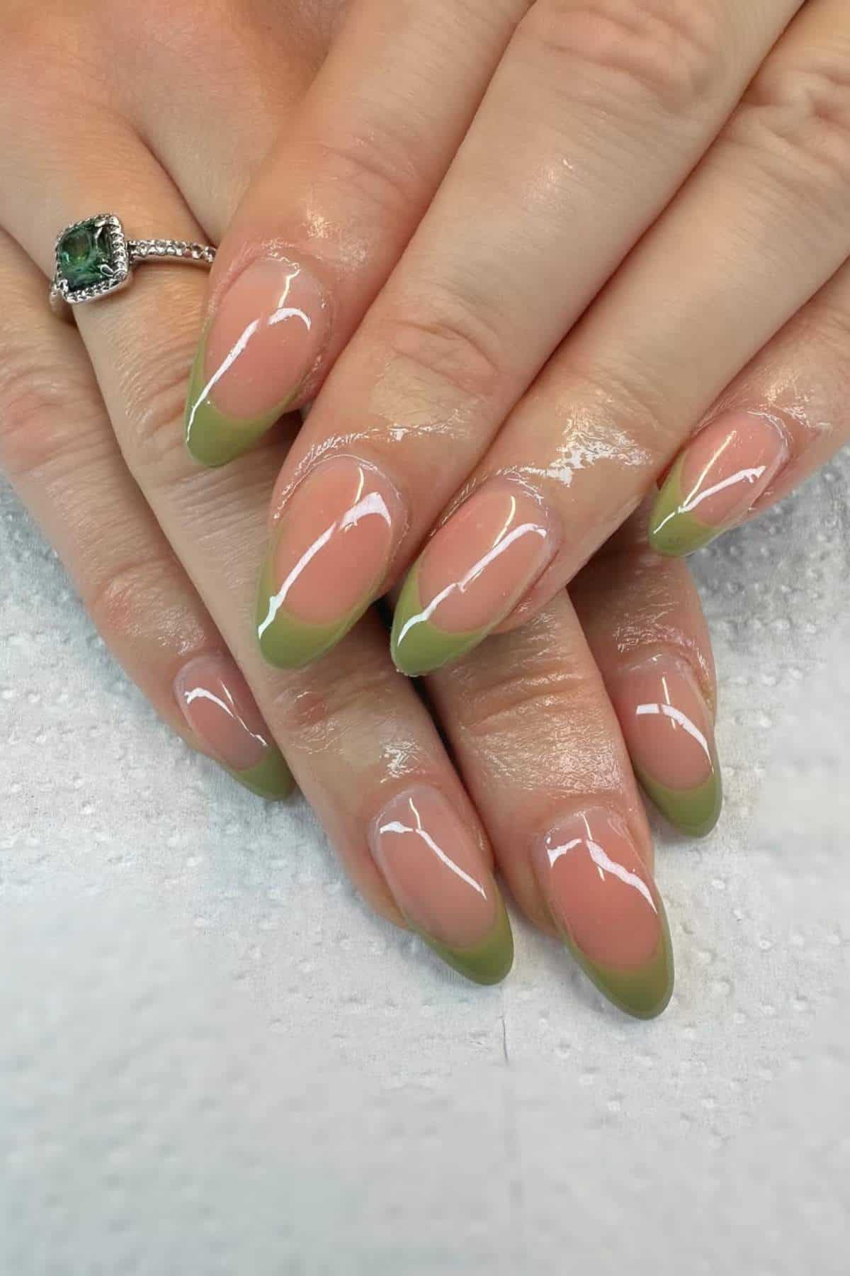 Hands with almond-shaped nails featuring a glossy nude base and green French tips. One hand has a silver ring with a large green gemstone.