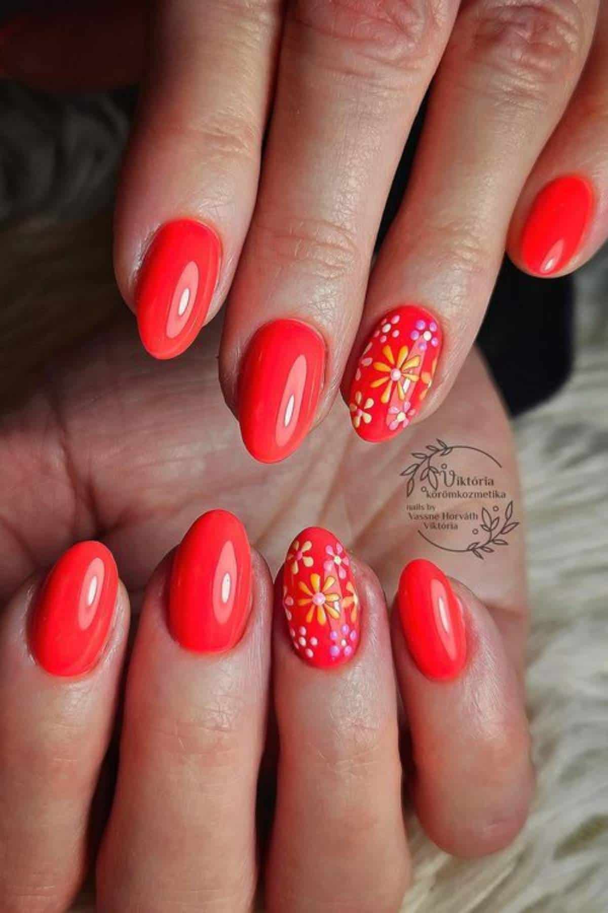 Hands with short, oval-shaped coral red nails; two accent nails feature yellow and orange floral designs with white dotted details.