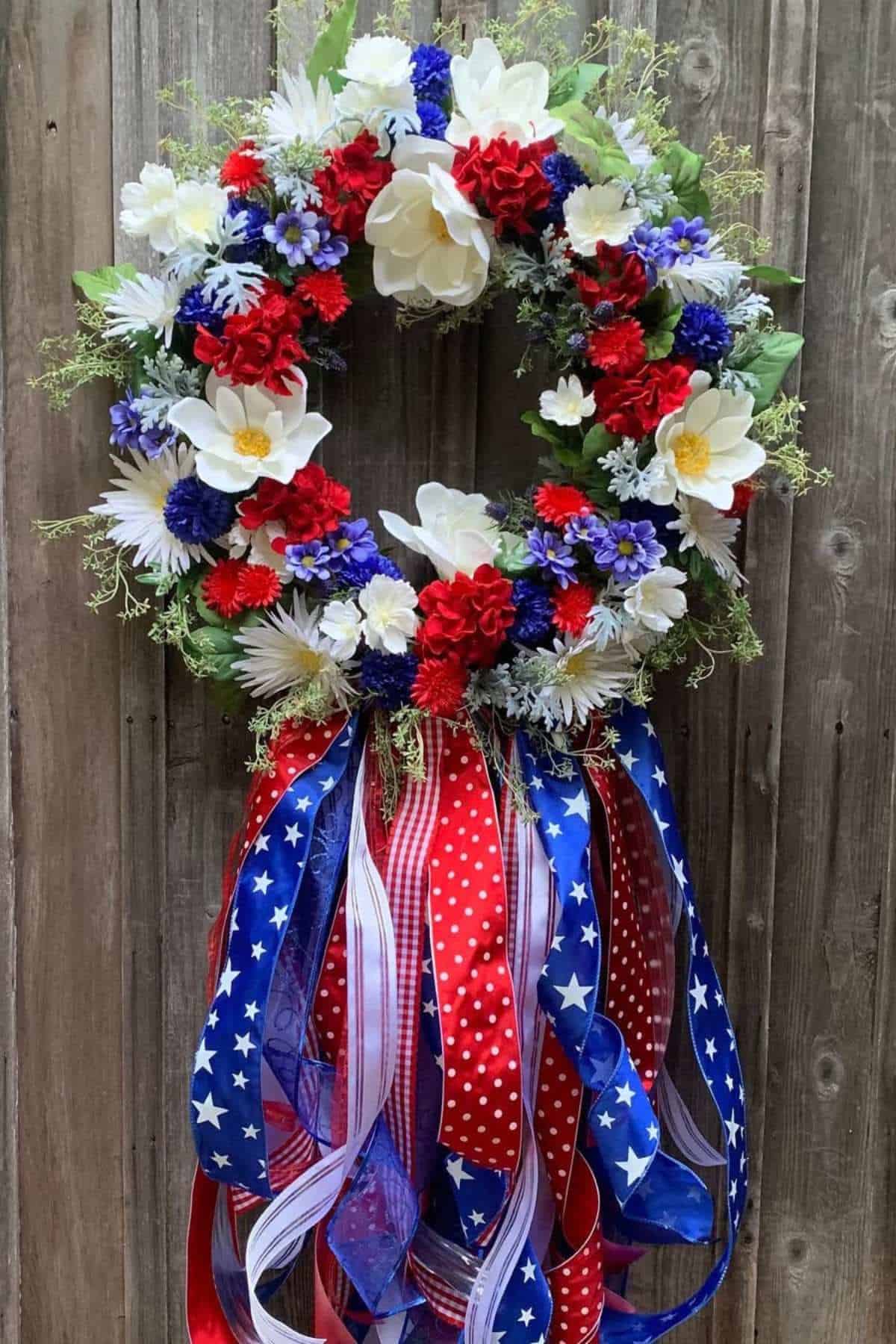 Patriotic wreath with red, white, and blue flowers and assorted ribbons featuring stars and stripes, hanging on a wooden wall.