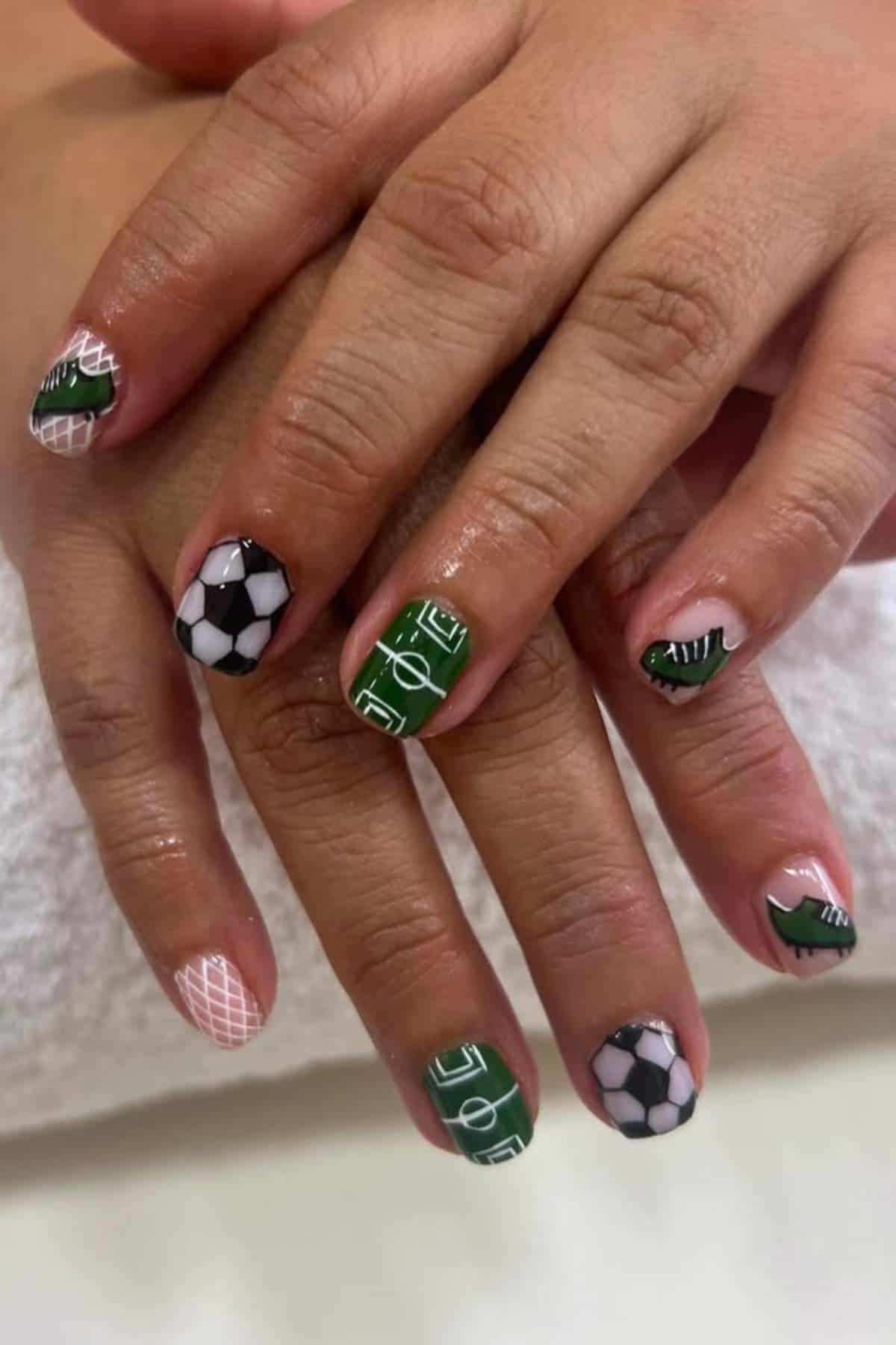 Close-up of hands with nail art featuring soccer balls, a soccer field, goal nets, and green and white designs on fingernails.