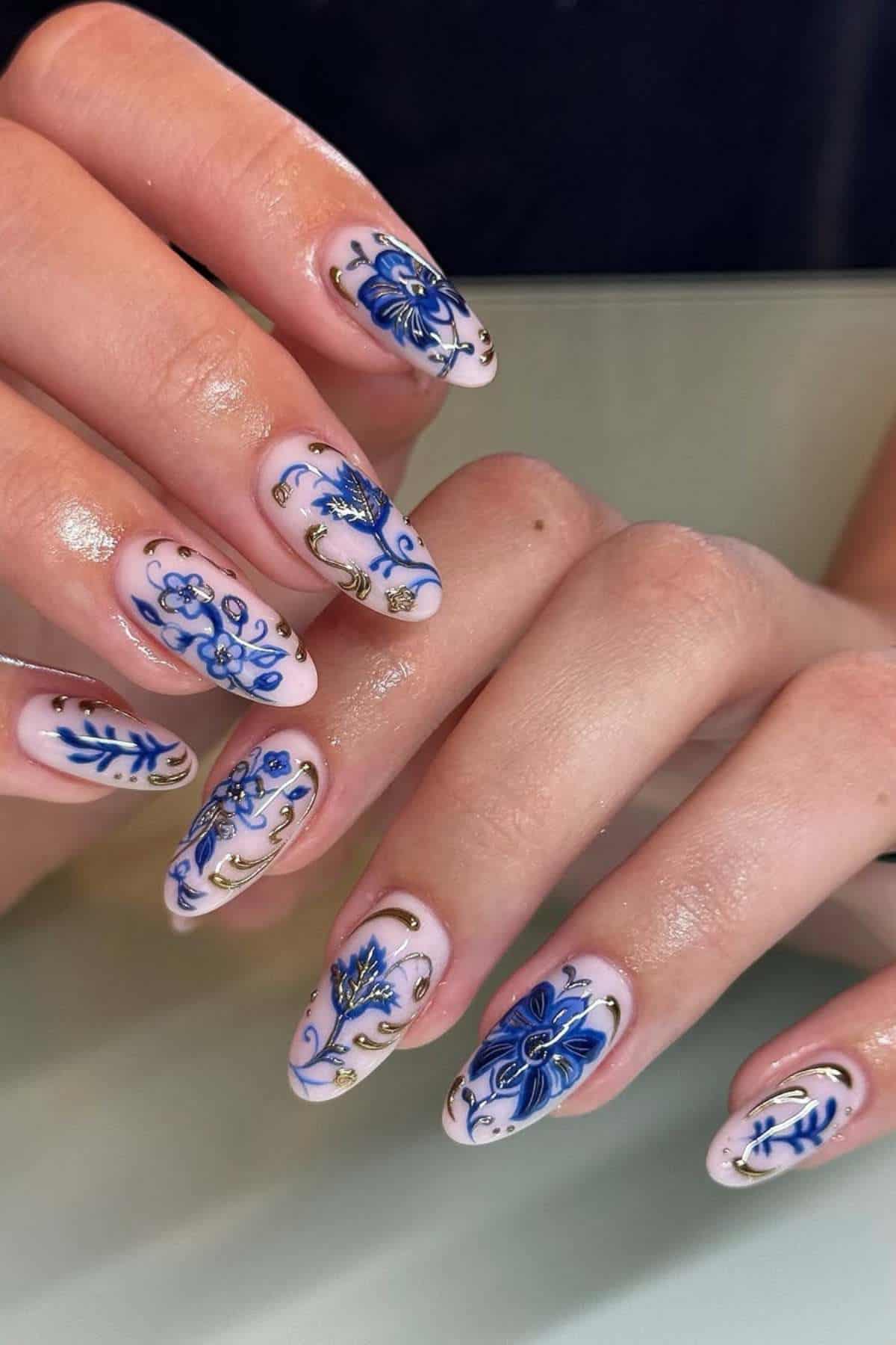 Hands with almond-shaped nails featuring intricate blue floral designs and gold accents on a pale pink base.